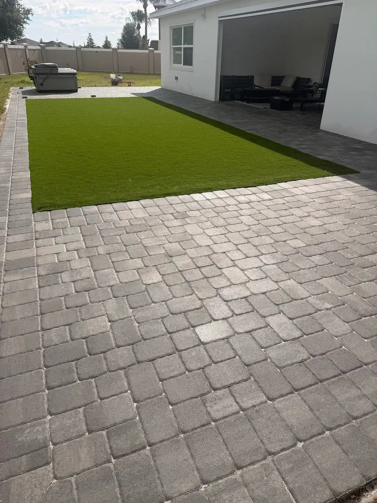 Rectangular green artificial turf lawn bordered by gray paver patio and adjacent to a white house patio area with outdoor furniture