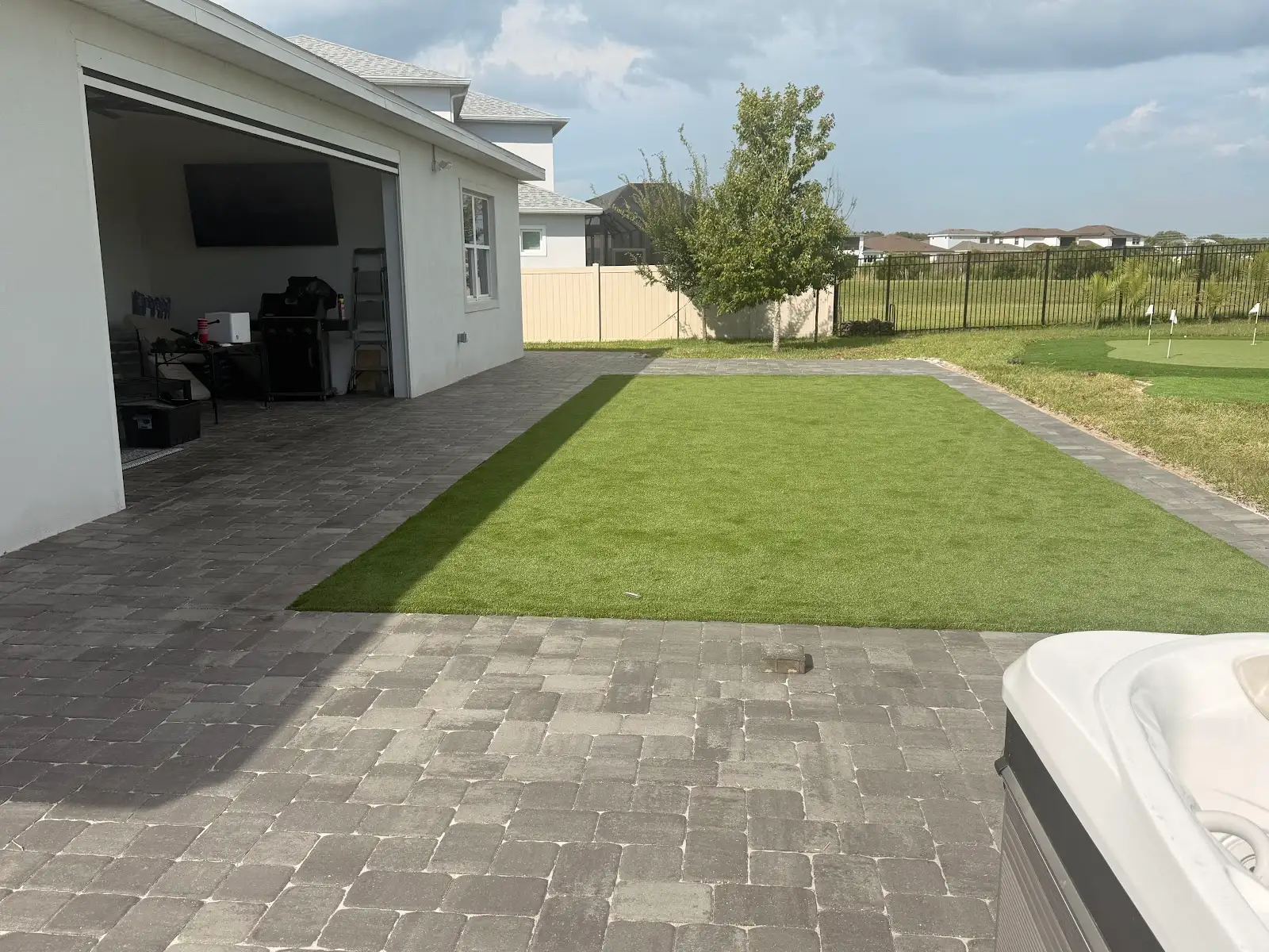 Backyard with a gray paver patio surrounding a rectangular area of artificial turf next to a white house and garage under a partly cloudy sky.