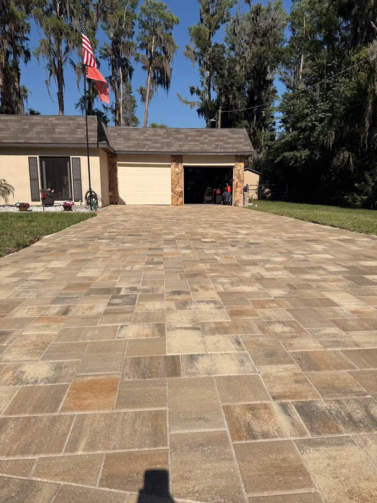 Wide view of a newly installed tan and brown rectangular paver driveway leading up to a two-car garage at a residential home in Tampa Bay, with trees and clear sky behind.