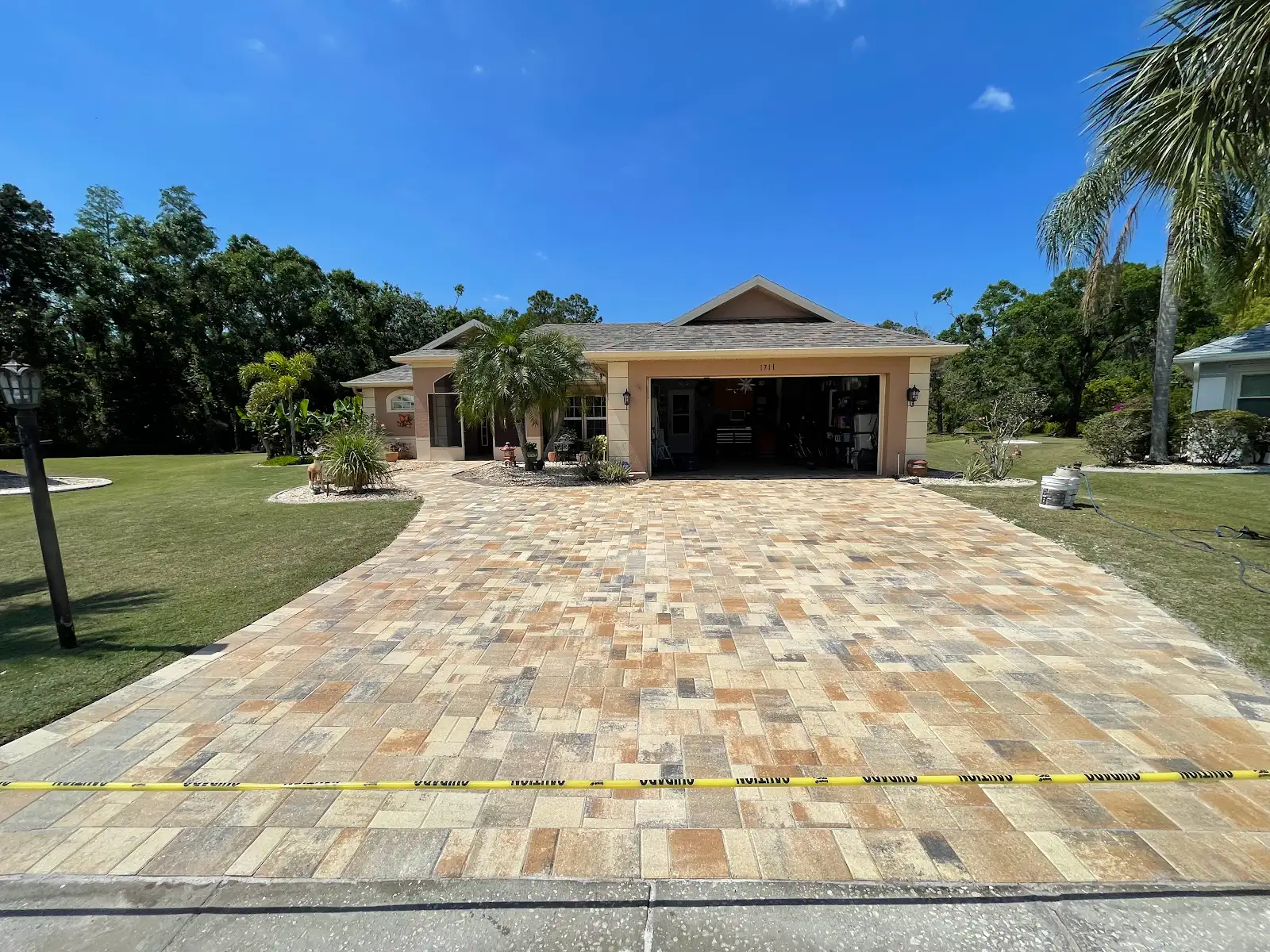 Stone paver driveway in front of a house with caution tape at the edge, surrounded by green lawn and palm trees under a blue sky.