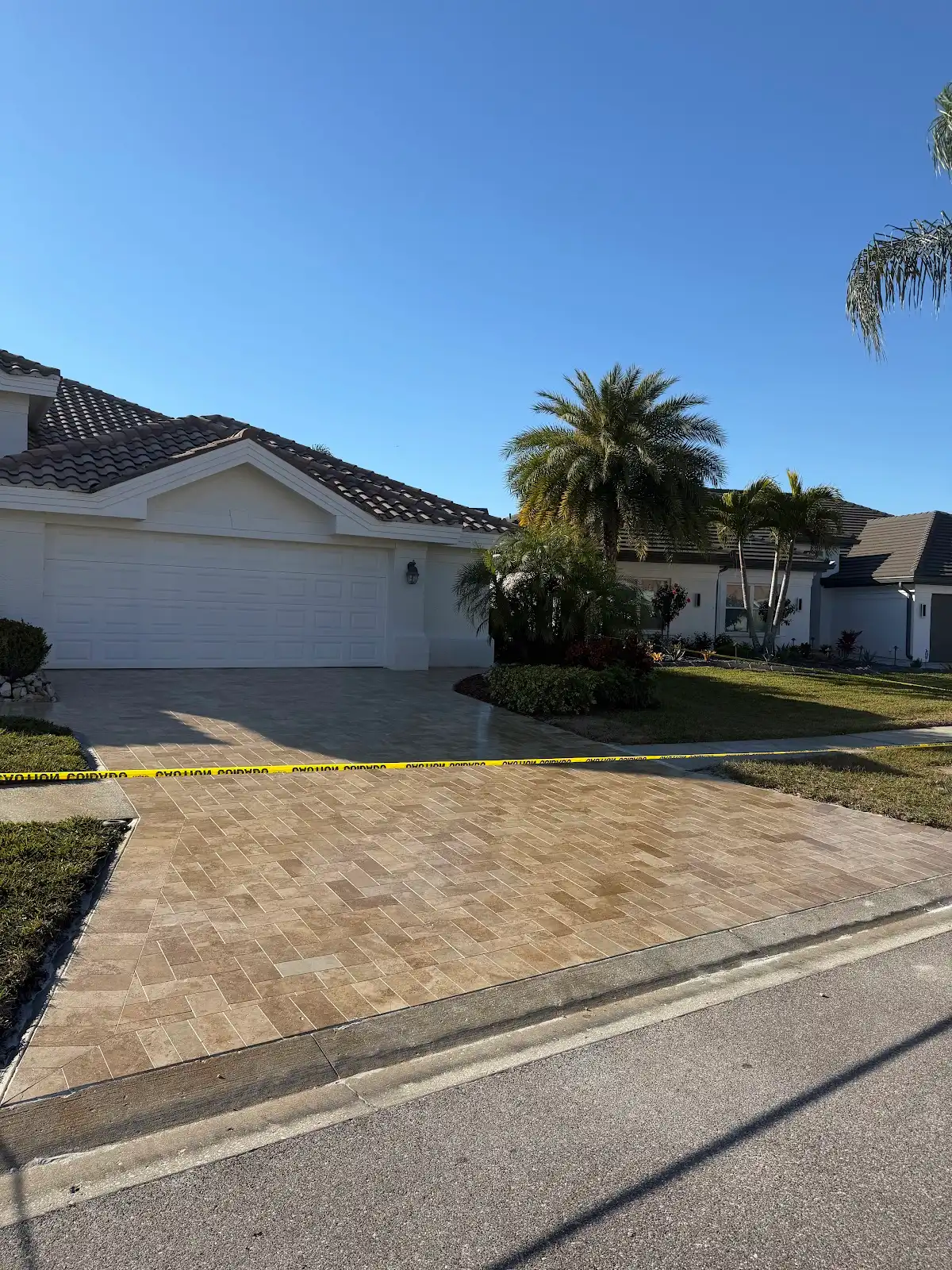 Freshly installed herringbone pattern paver driveway in front of a white garage with palm trees and landscaping on the side, under a clear blue sky.