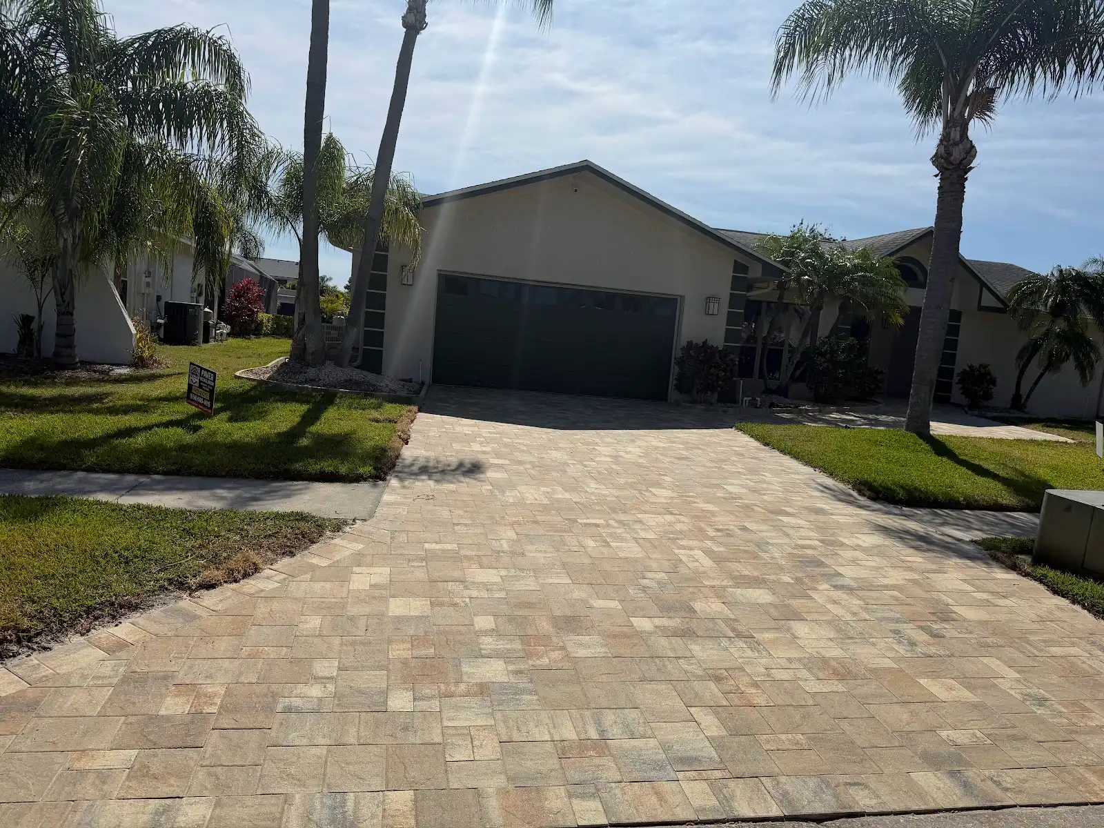 Residential driveway paved with beige and light brown pavers leading to a two-car garage with green grass and palm trees nearby.