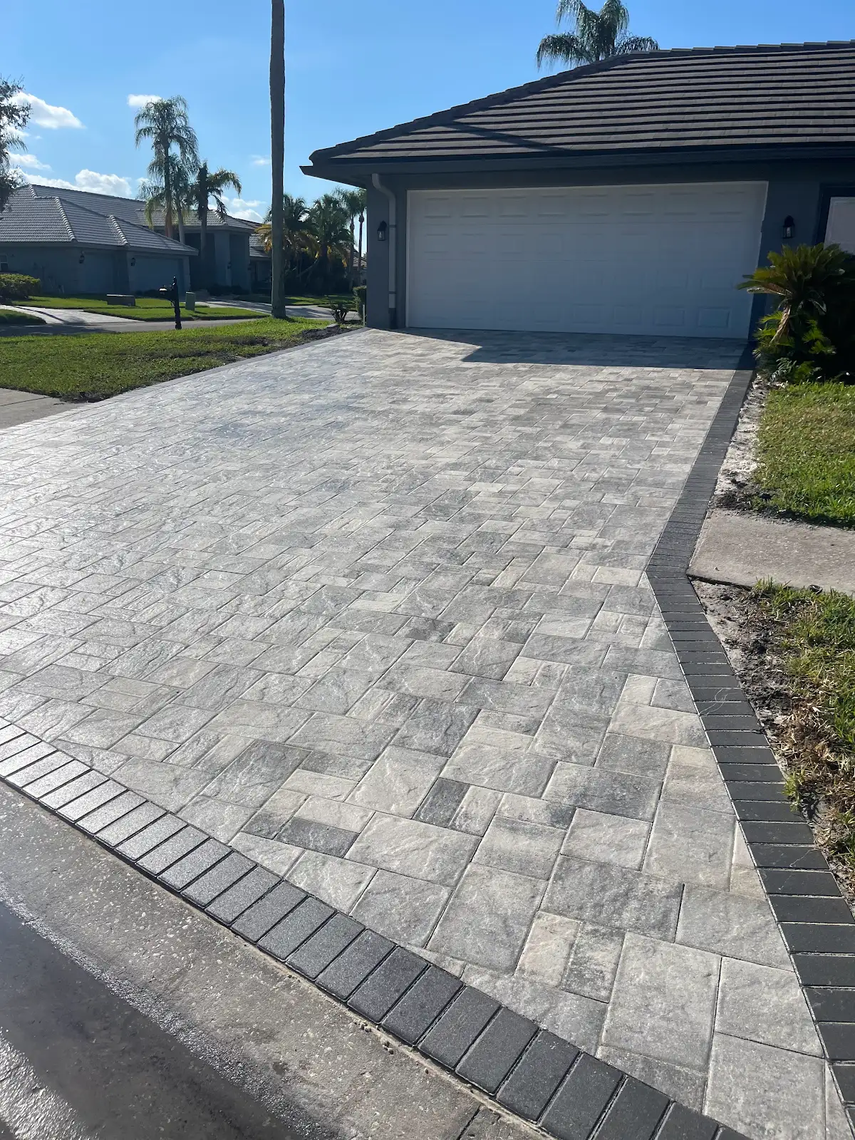 Light gray patterned paver driveway bordered by dark gray pavers leading up to a white two-car garage door during daytime in Tampa Bay.