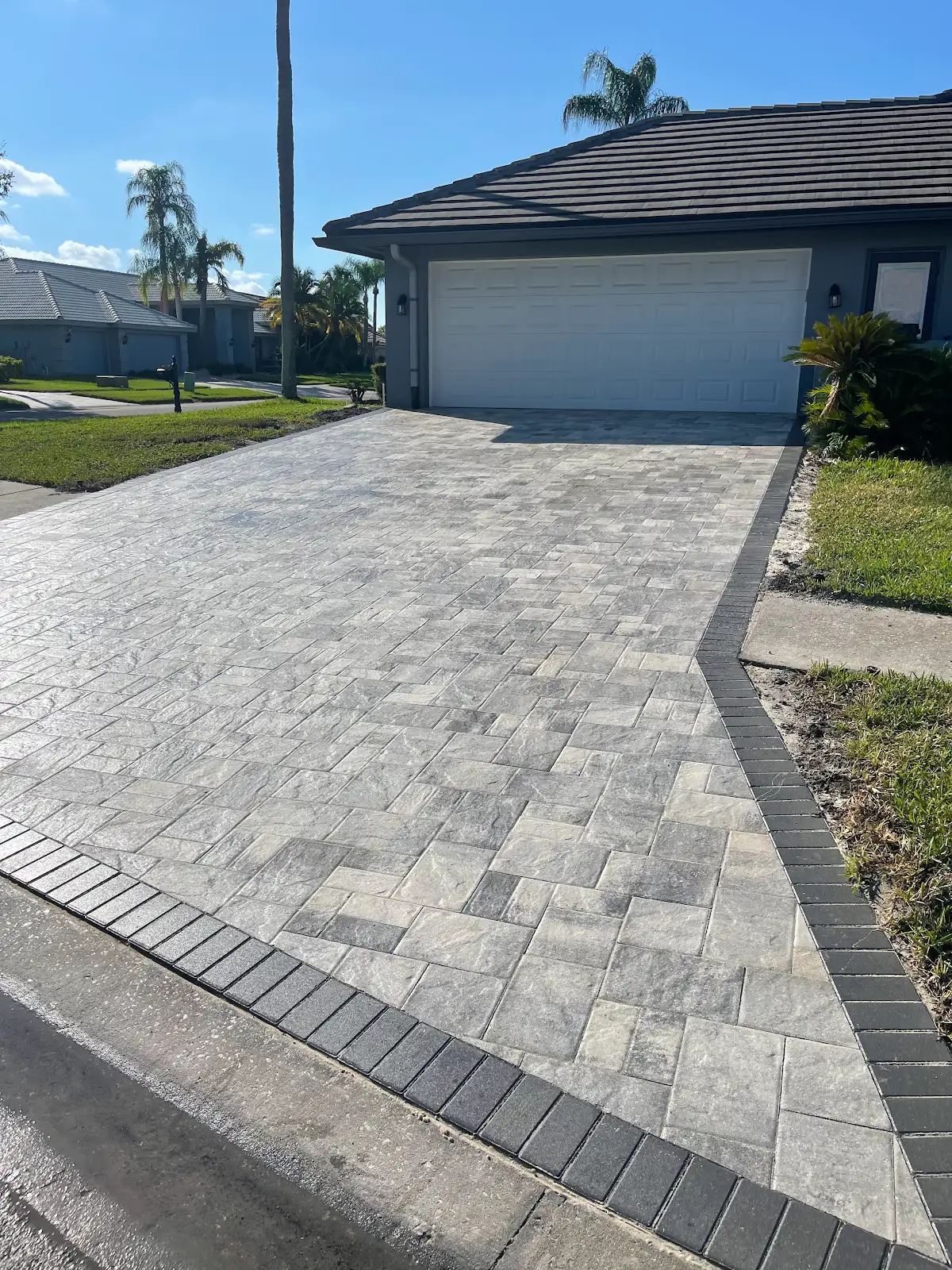 Residential driveway paved with grey interlocking pavers and a dark brick border under clear blue sky in Tampa Bay