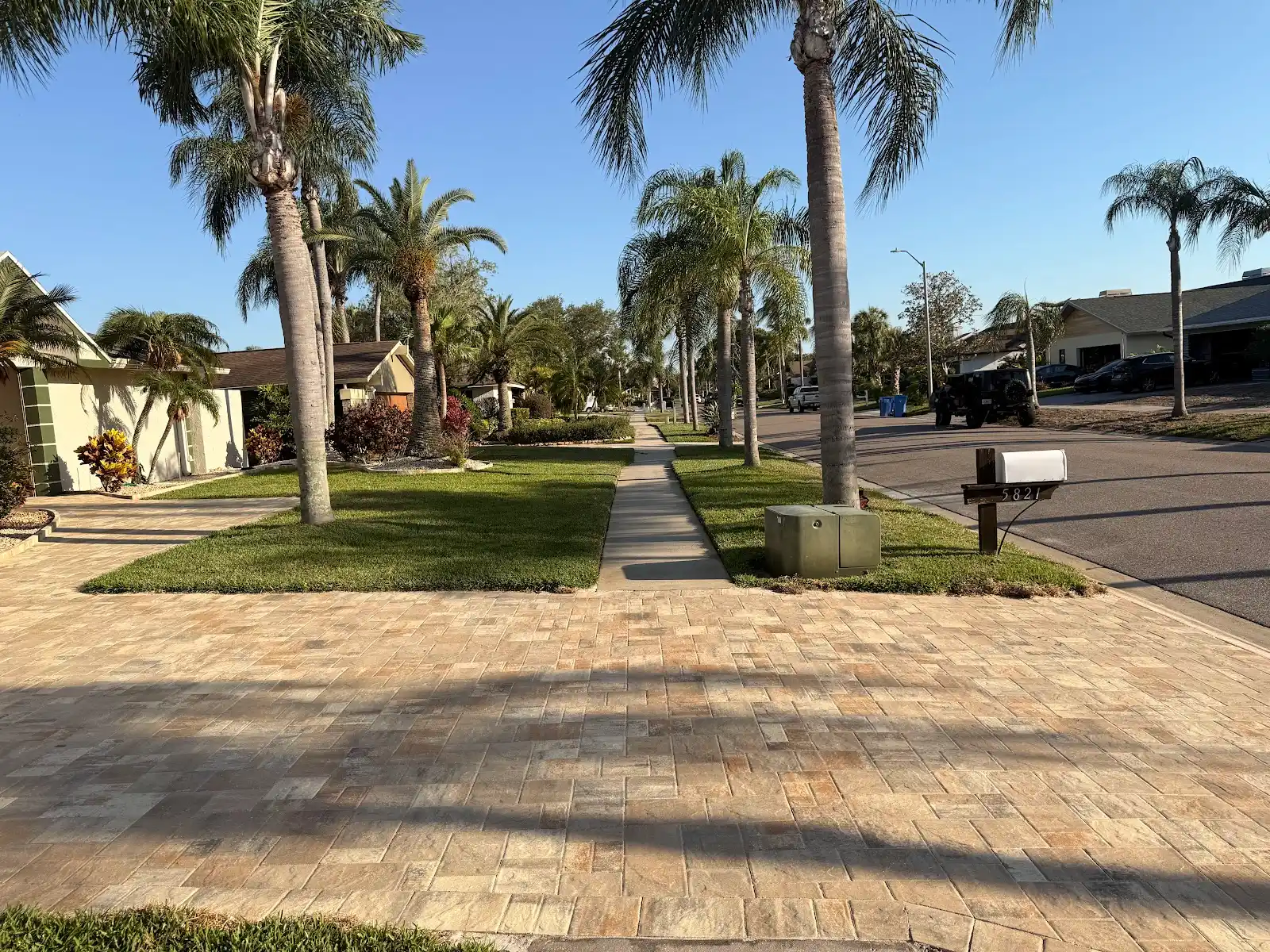 Light tan rectangular paver driveway leading to a house, with palm trees and grass lawns on both sides under a clear sky.