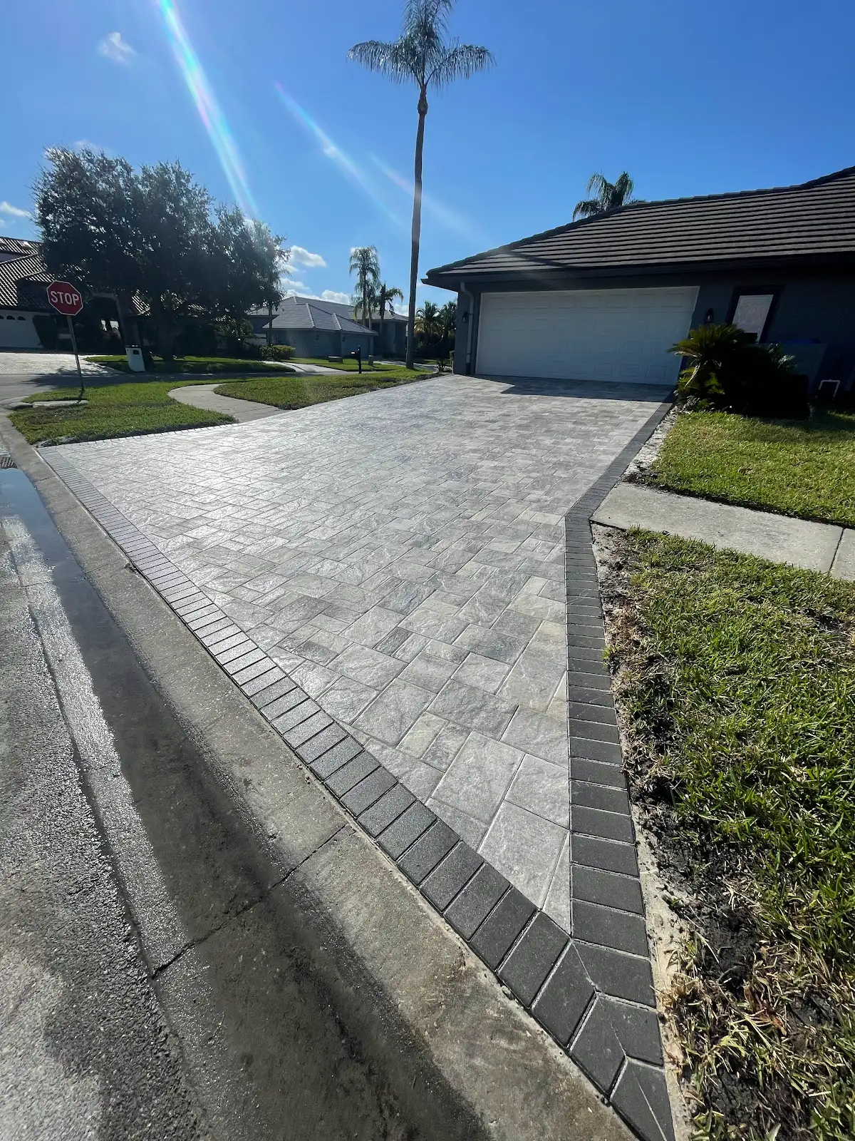 Driveway made of light gray pavers with dark gray border bricks leading to a garage of a house with palm trees and grass on sides.
