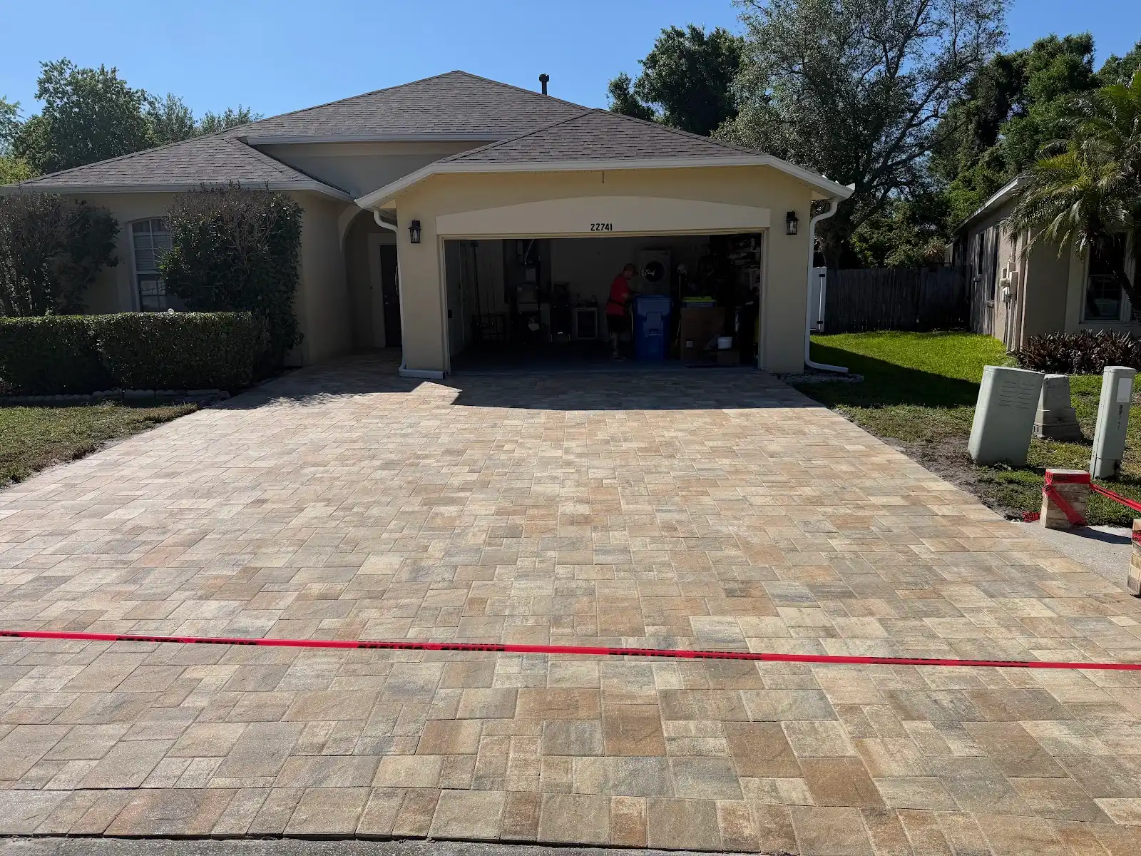 Wide view of a new multi-tone paver driveway leading up to a beige residential garage with the garage door open and a red caution tape stretched across the driveway entrance.