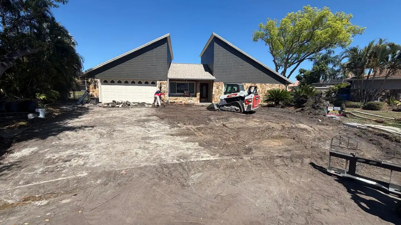 Construction site with cleared driveway in front of house, worker and small loader leveling ground under clear blue sky