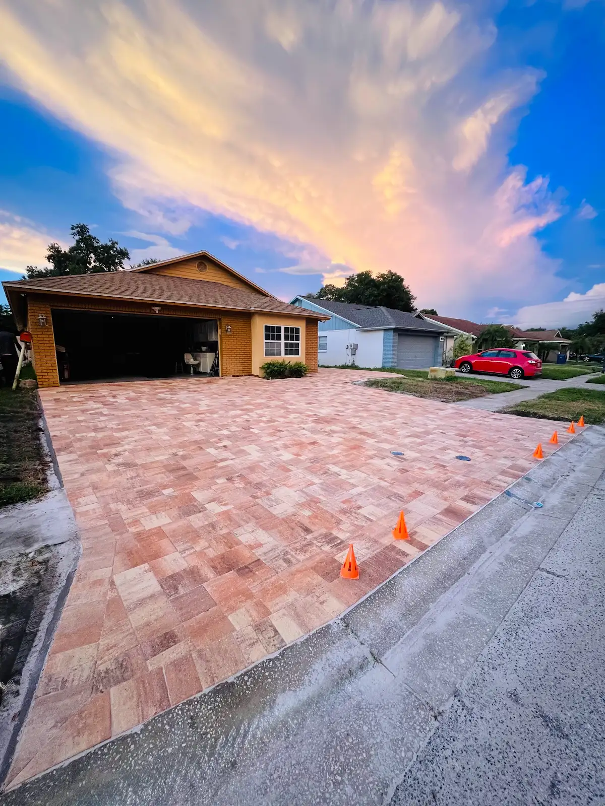 Wide view of a newly installed brick paver driveway in front of a single-story house at sunset with orange cones marking its edge.
