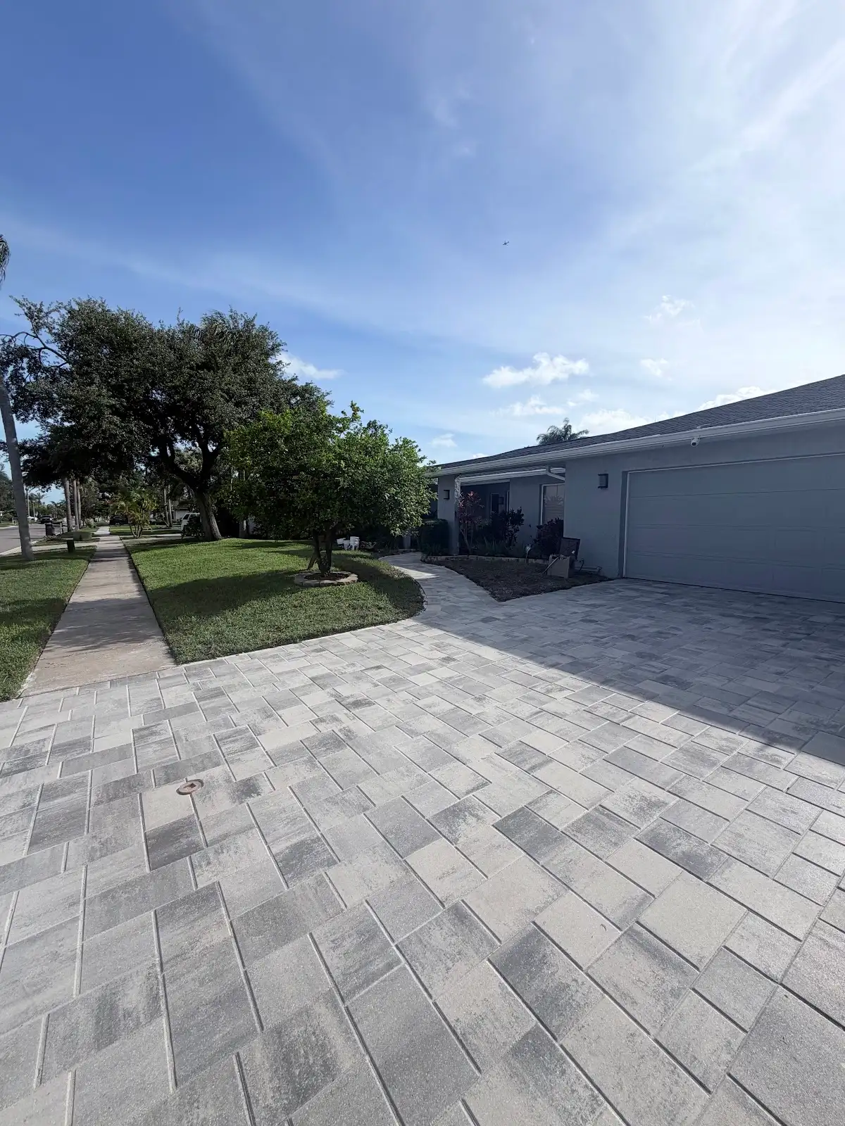 Light gray interlocking paver driveway and curved walkway beside a house garage with green lawn and trees under blue sky