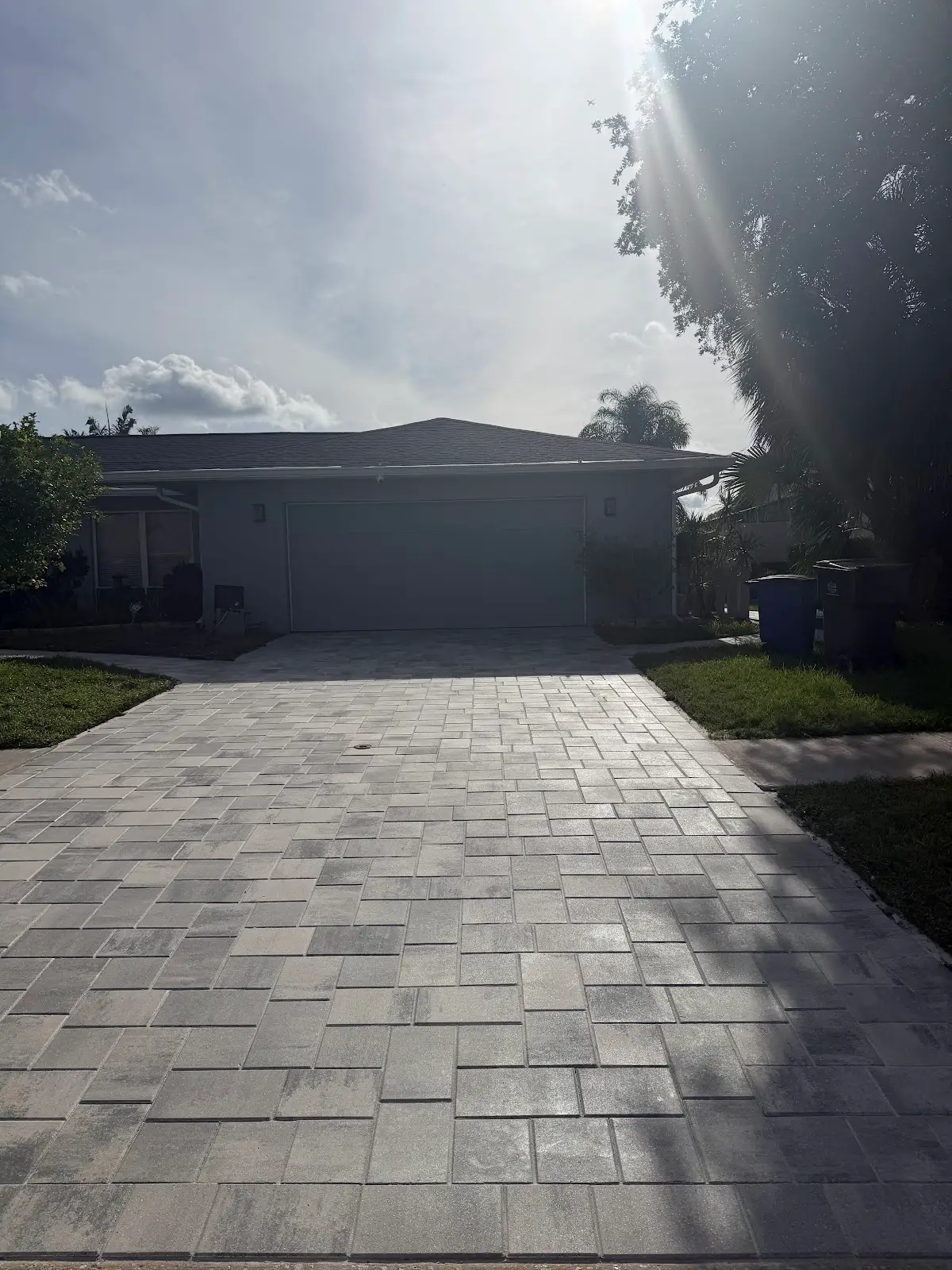 Light gray paver driveway leading to a garage in a residential home with some grass and trees on the sides under a bright sky.