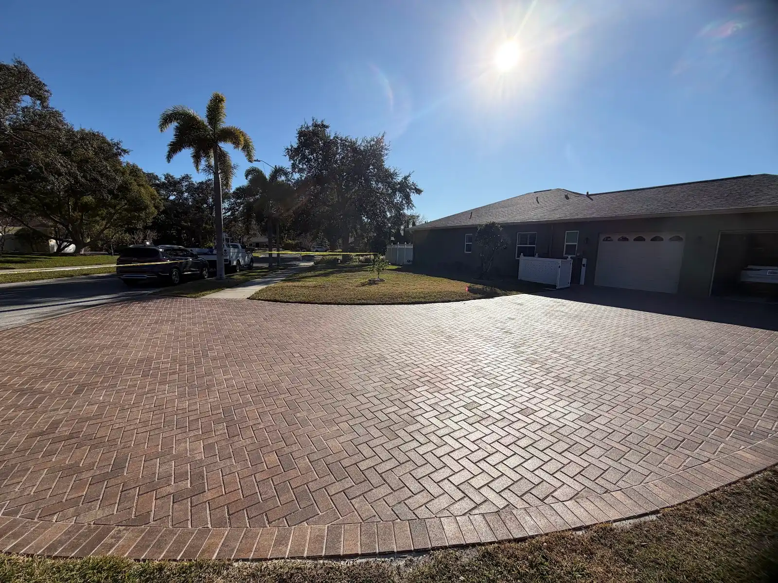 Wide view of a newly installed reddish-brown paver driveway in front of a house with adjacent lawn and trees under a bright sun