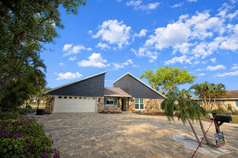 Wide view of a large paver driveway leading to a two-car garage of a house with stone and dark siding, with trees and bushes on sides under a partly cloudy sky.
