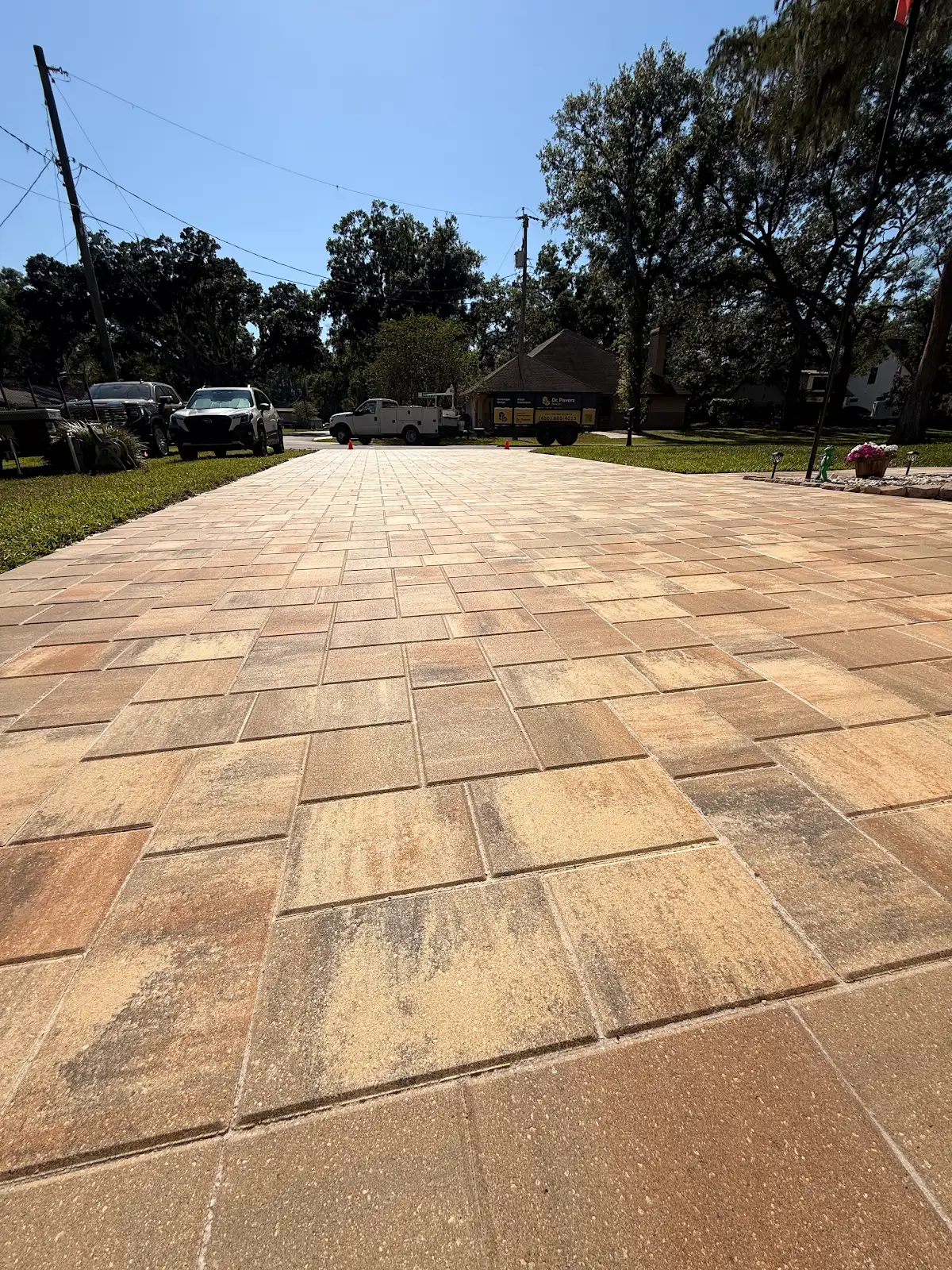 Close-up view of a freshly installed beige and brown paver driveway with rectangular stones and a clear blue sky, surrounded by grass and trees.