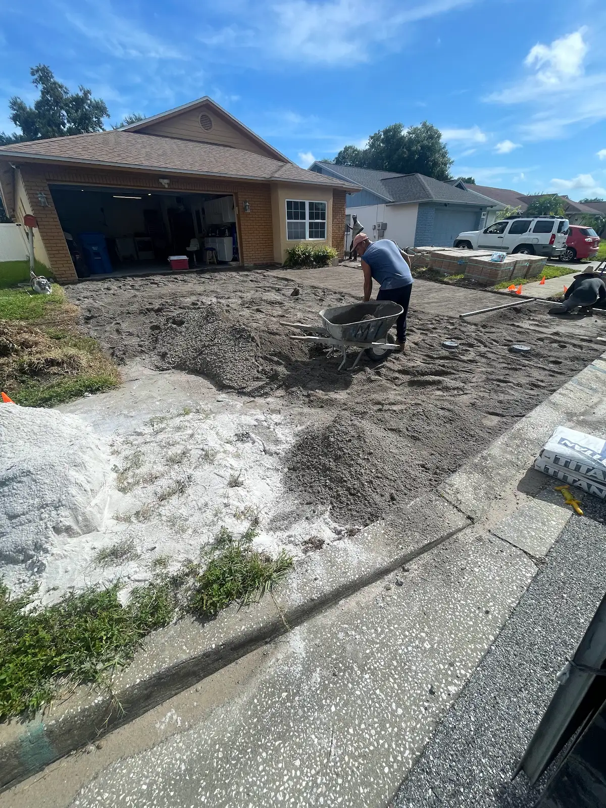Worker preparing ground and base materials for driveway paver installation in front of a brick house on a sunny day
