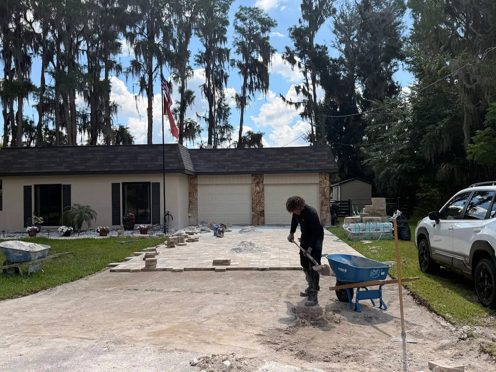 Worker leveling ground for paver driveway with partially laid pavers in front of a two-car garage and a house with tall trees in background