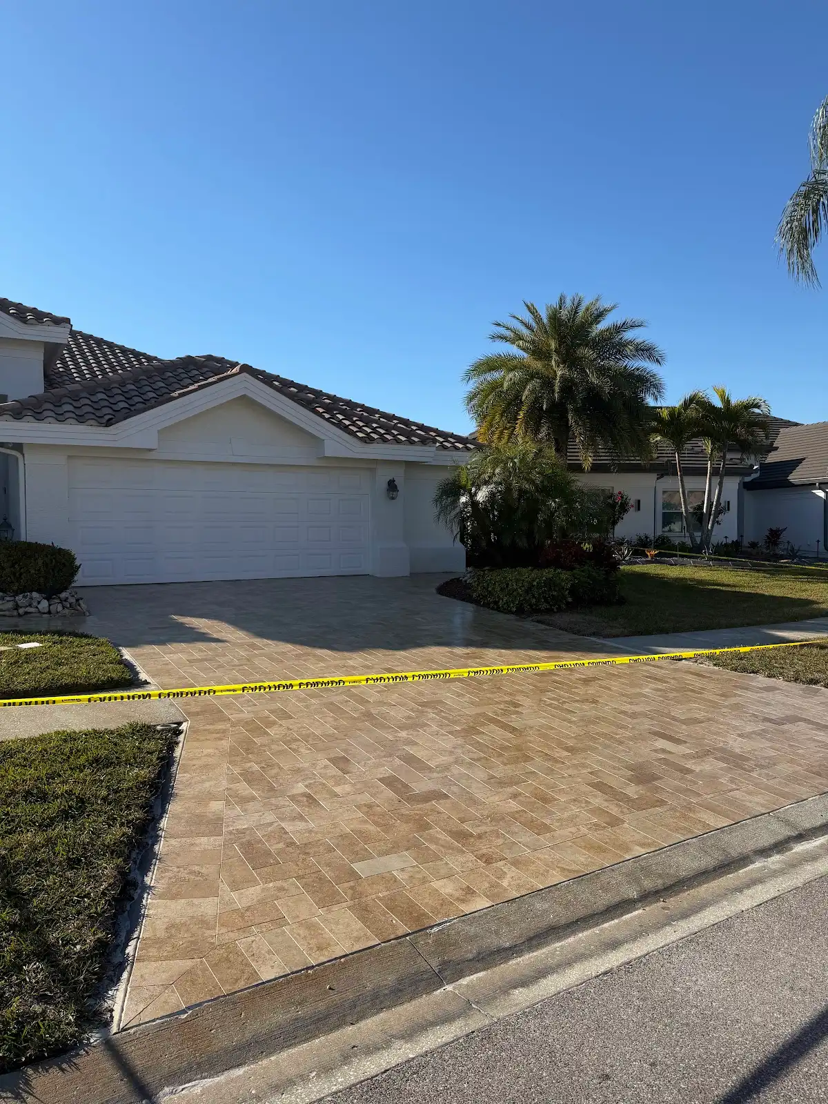 Light tan herringbone patterned paver driveway in front of a white house with tiled roof, surrounded by greenery and yellow caution tape.