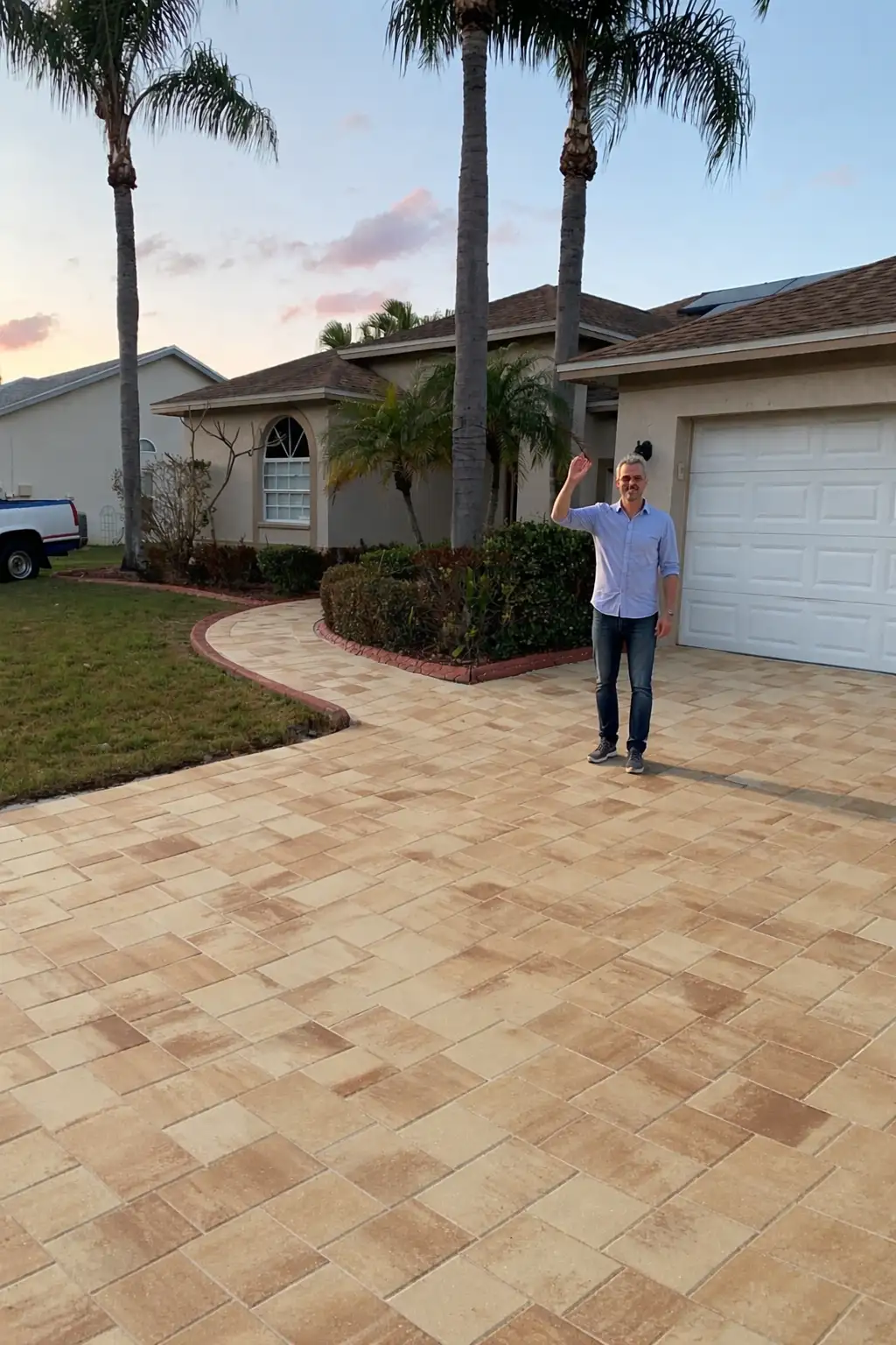 Beige and light brown paver driveway and curved walkway outside a house with palm trees, a man waving on the driveway under evening sky
