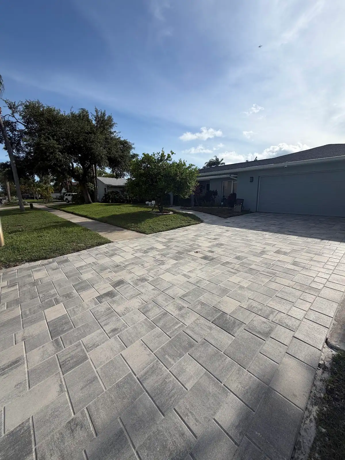 Light grey and beige paver driveway in front of a house with garage, lawn, and trees on a sunny day