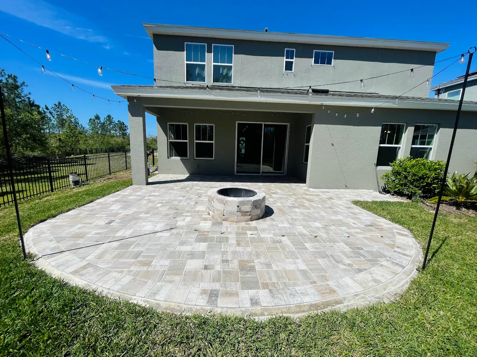 A backyard patio paved with light-colored bricks featuring a circular firepit in the center, next to a gray two-story house with a covered porch and string lights above.