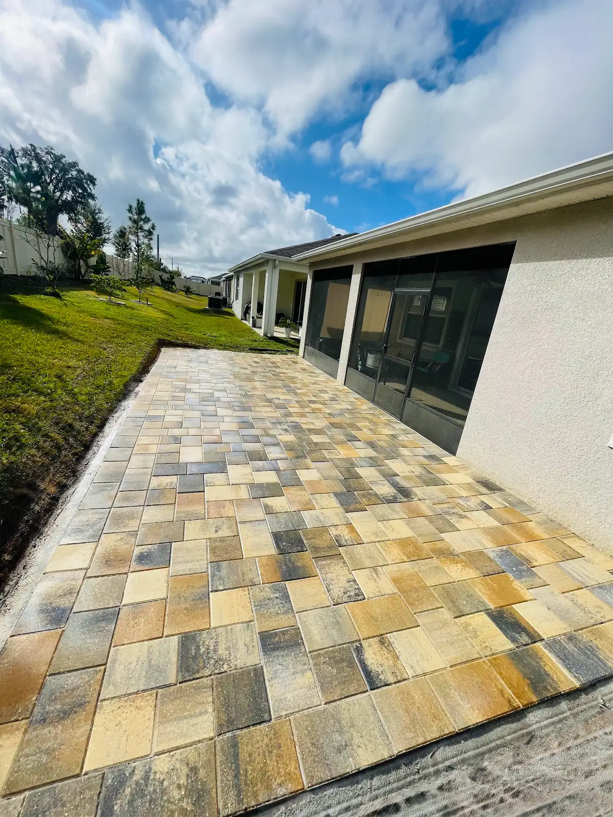 Rectangular multi-tone paver patio next to screened porch and green lawn under partly cloudy sky
