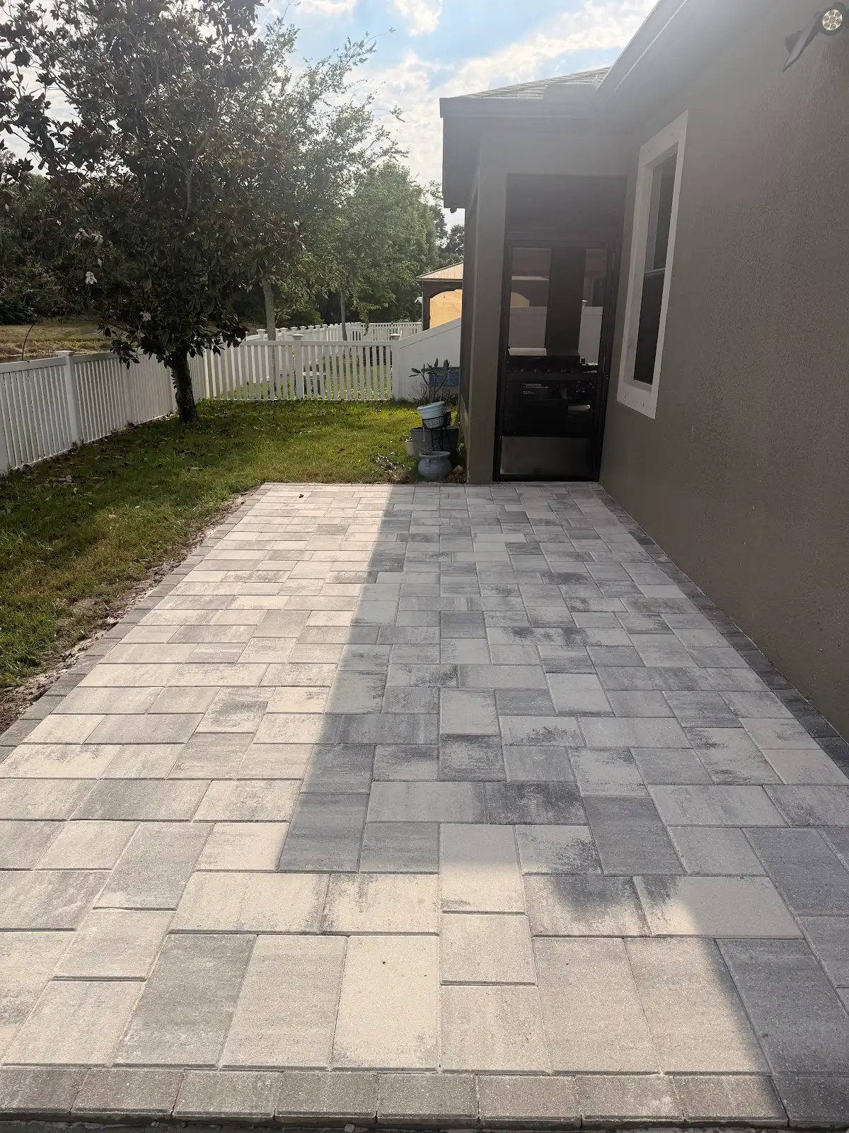 Gray rectangular paver patio next to a house with a tree and white fence in the background on a sunny day.