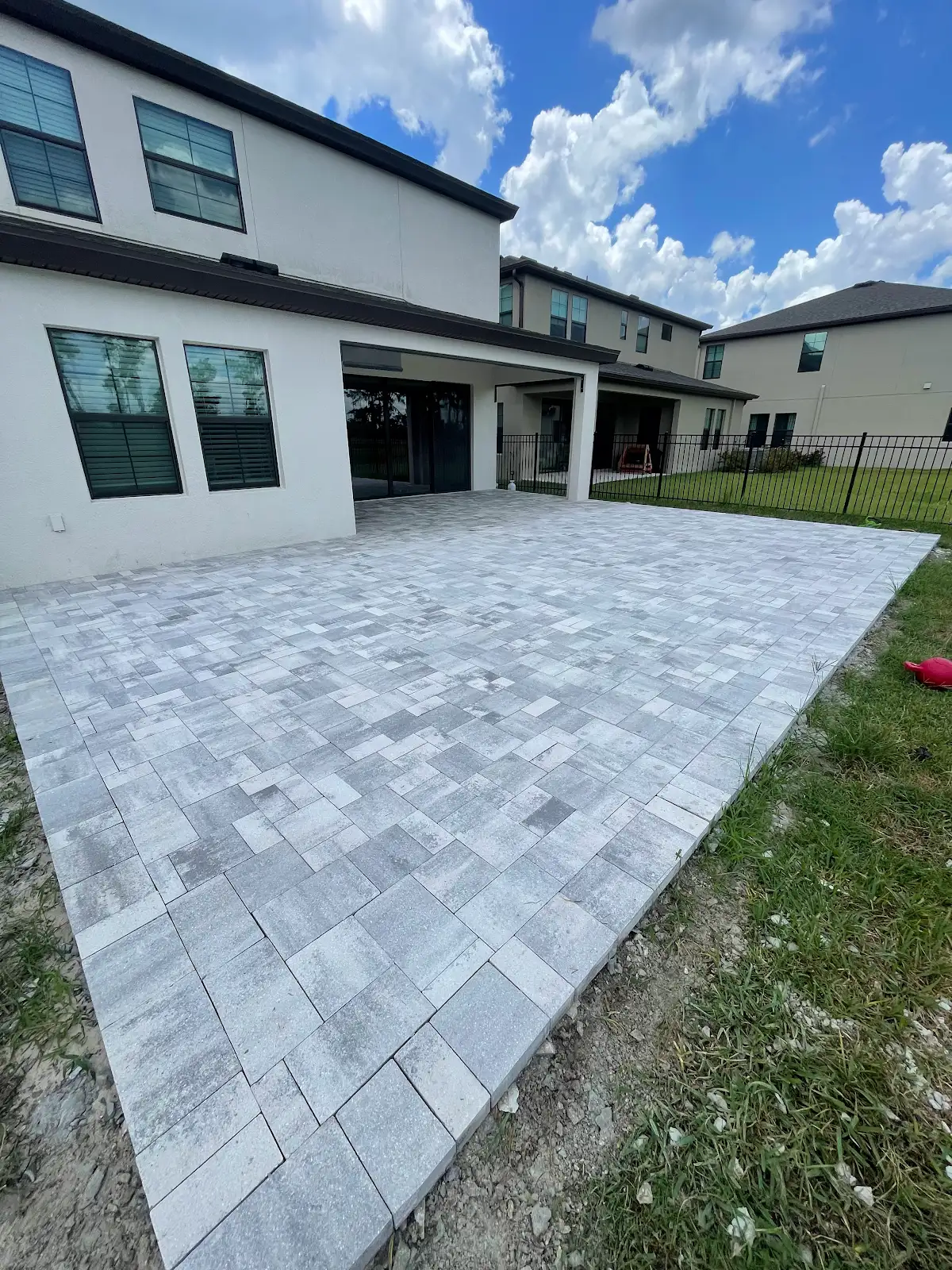 Gray and white rectangular paver patio in progress beside a white house with multiple windows and sliding glass doors, partly surrounded by grass and metal fencing under a partly cloudy sky.