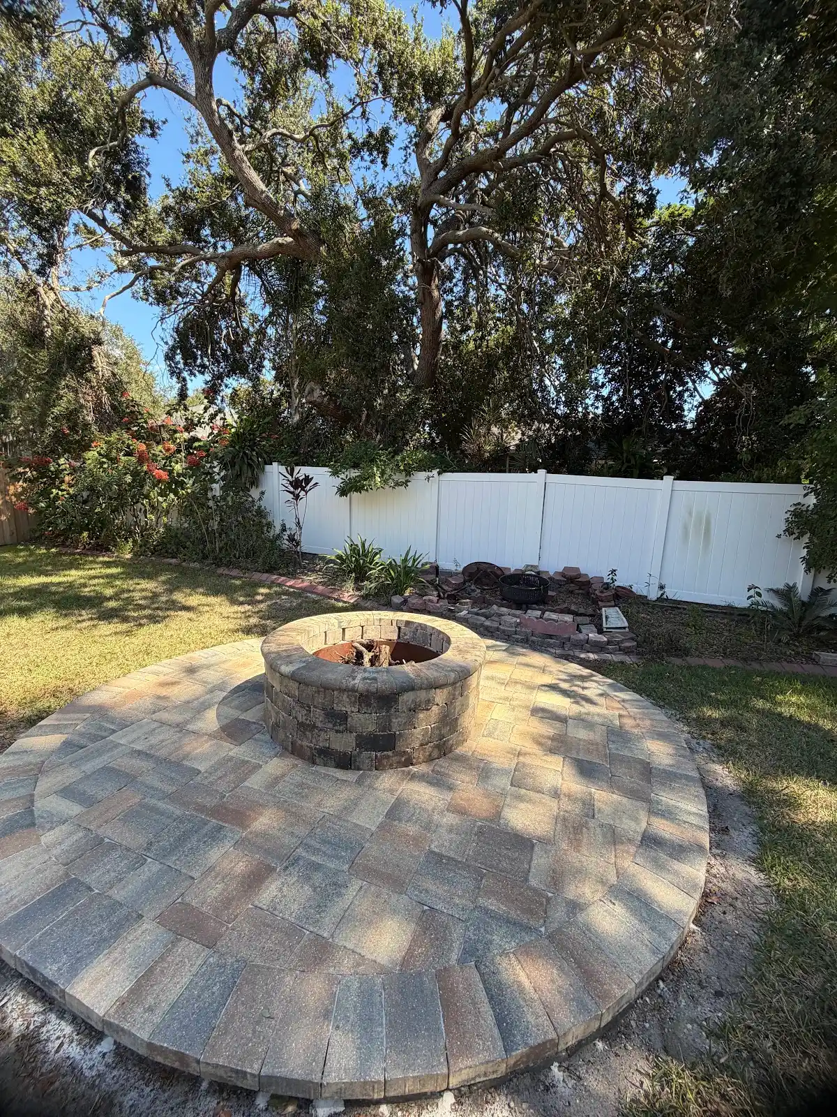 Circular paver patio with a brick fire pit in the center, surrounded by grass, plants, white fence and trees in the background under a clear sky.