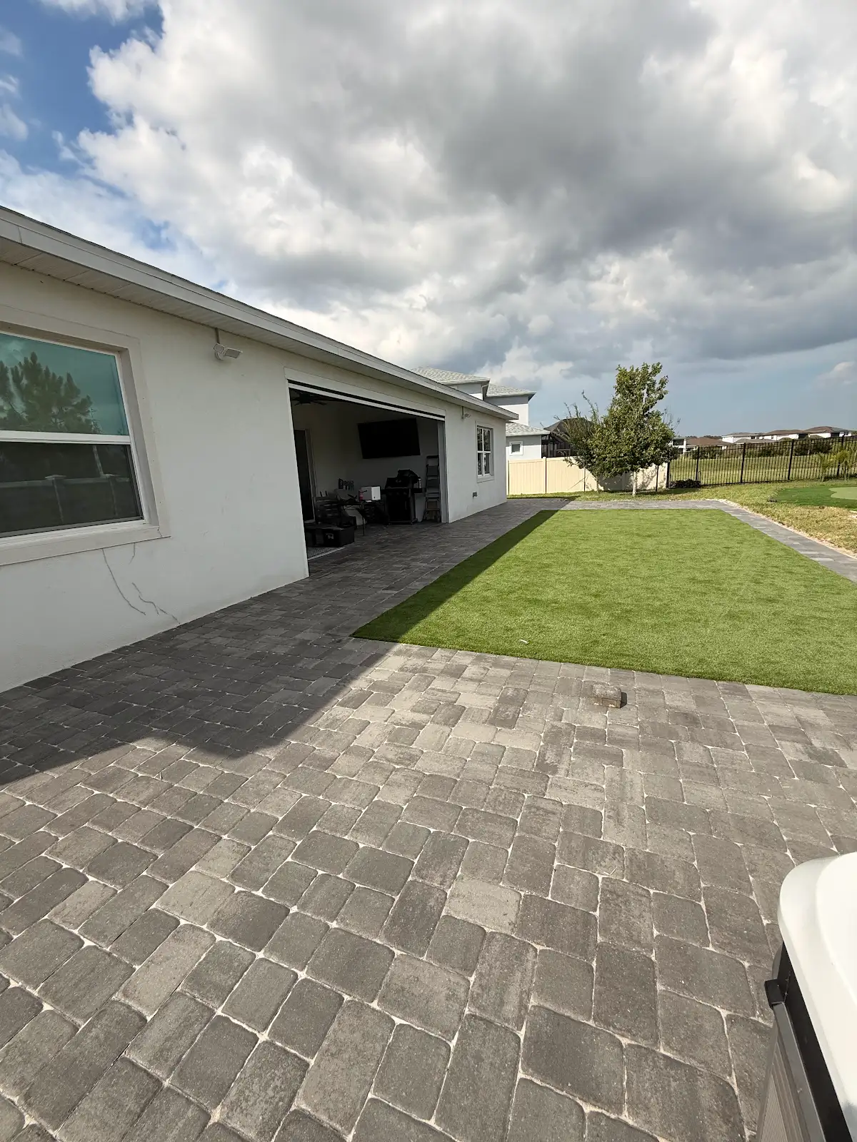 Backyard patio with gray paver stones and green artificial turf next to white house and cloudy sky