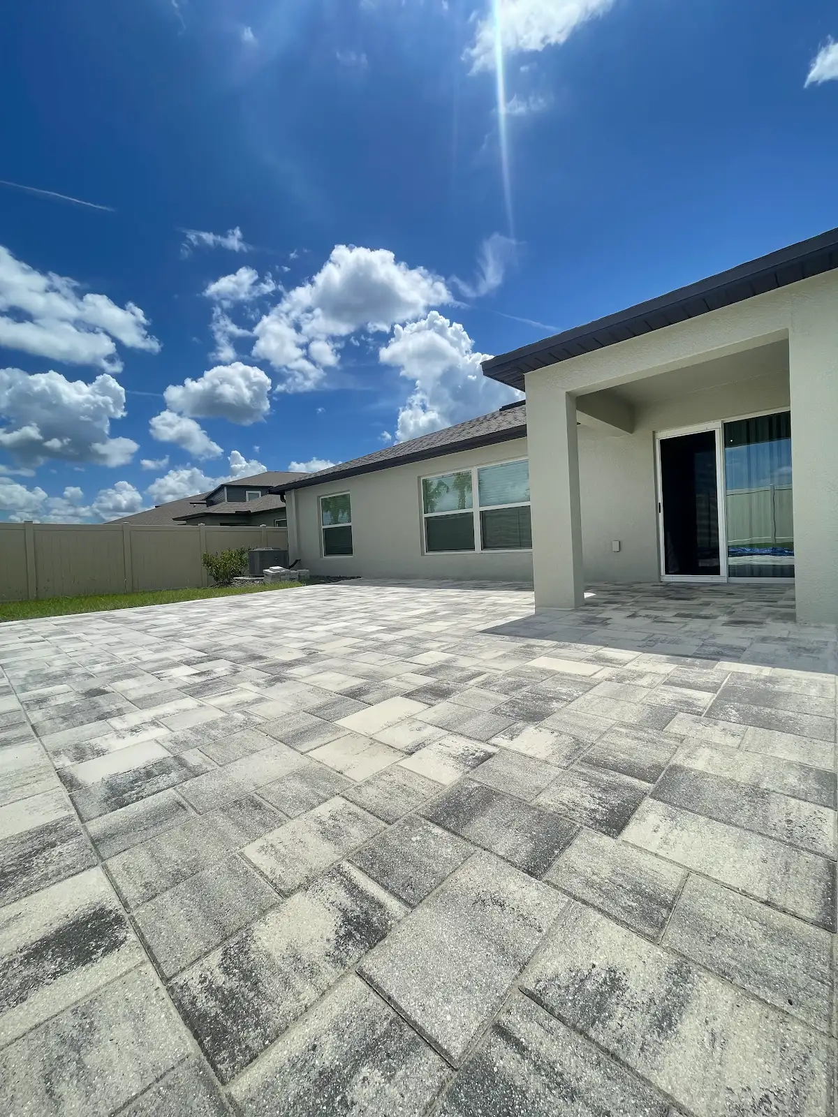 Large patio area paved with gray and white patterned pavers next to a house with sliding glass doors and covered patio, under sunny blue sky with clouds
