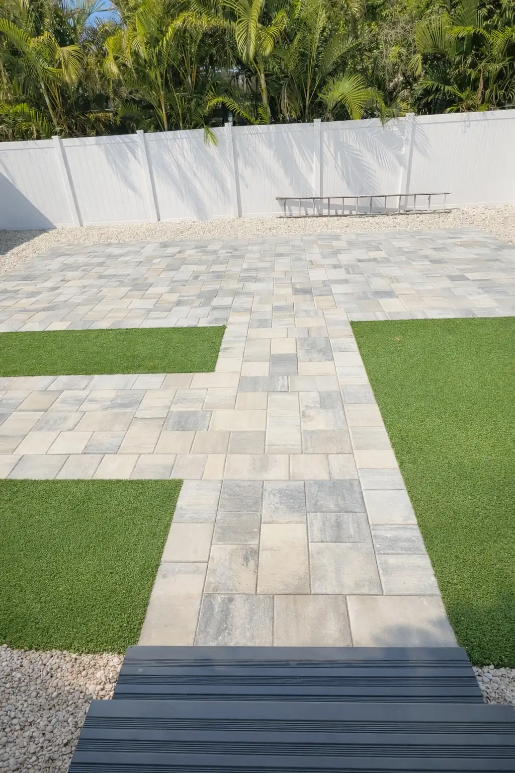 Outdoor patio area featuring light gray and beige pavers with artificial turf sections and white gravel, backed by a white fence and tropical greenery.