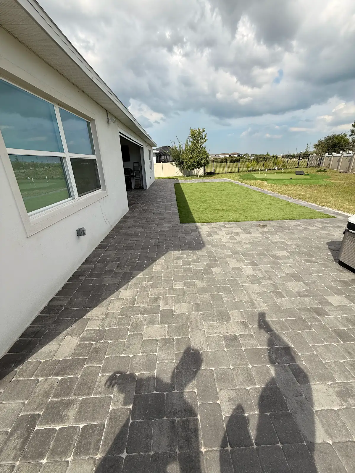 Grey stone paver patio next to a white house with a square patch of artificial turf and backyard putting green under partly cloudy sky