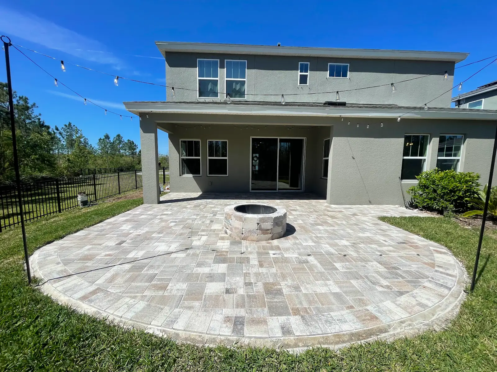 Curved light-colored paver patio with a circular stone fire pit in the center in backyard of two-story house, green lawn surrounding and string lights overhead on poles.