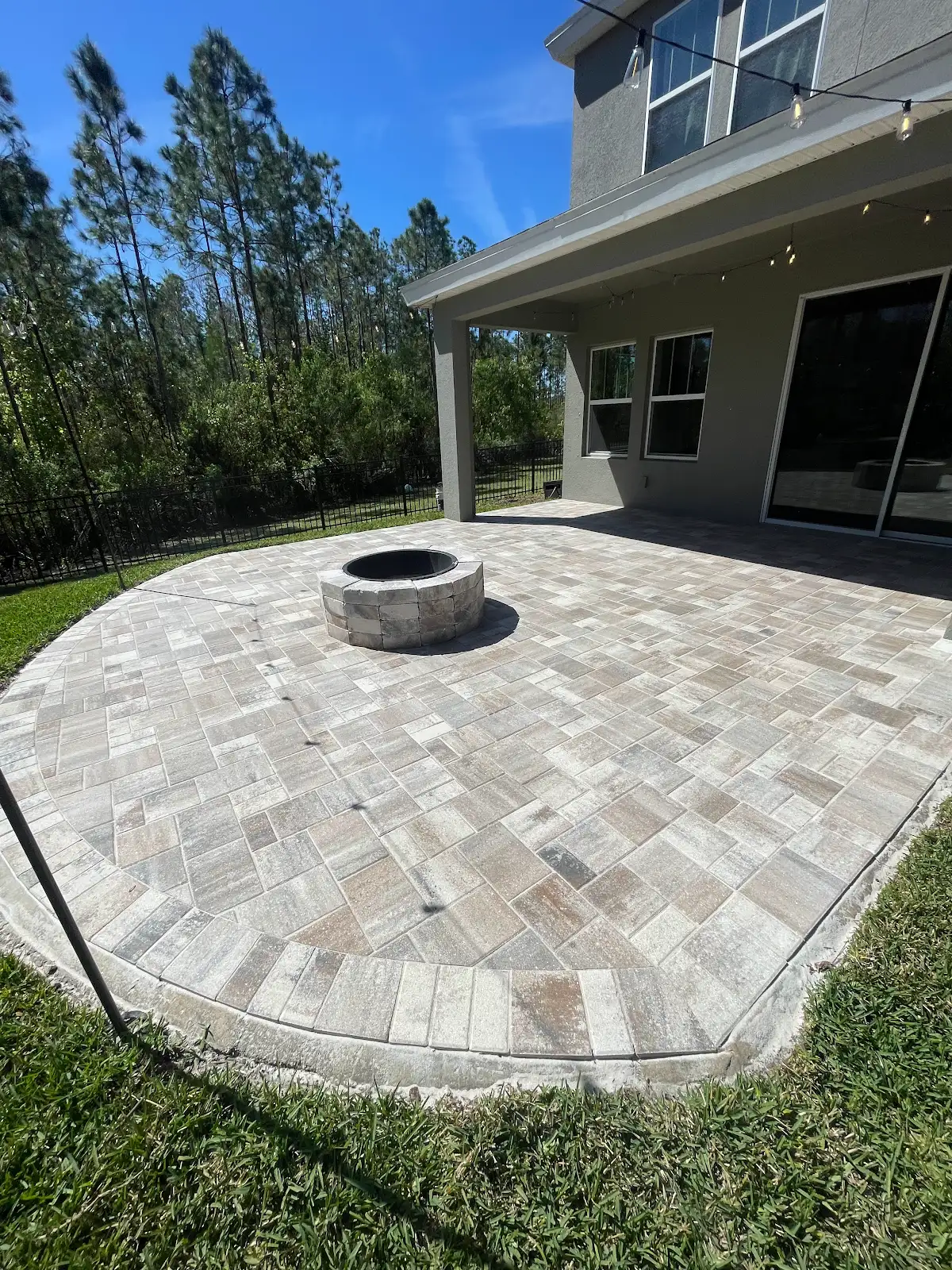 Outdoor paver patio with built-in circular fire pit, adjacent to a house, green grass surrounding the patio and trees in the background under a clear blue sky