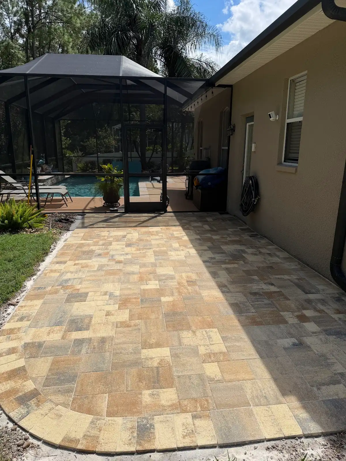 A freshly installed patterned paver patio leading to a screened pool enclosure next to a beige house wall in a sunny backyard setting.