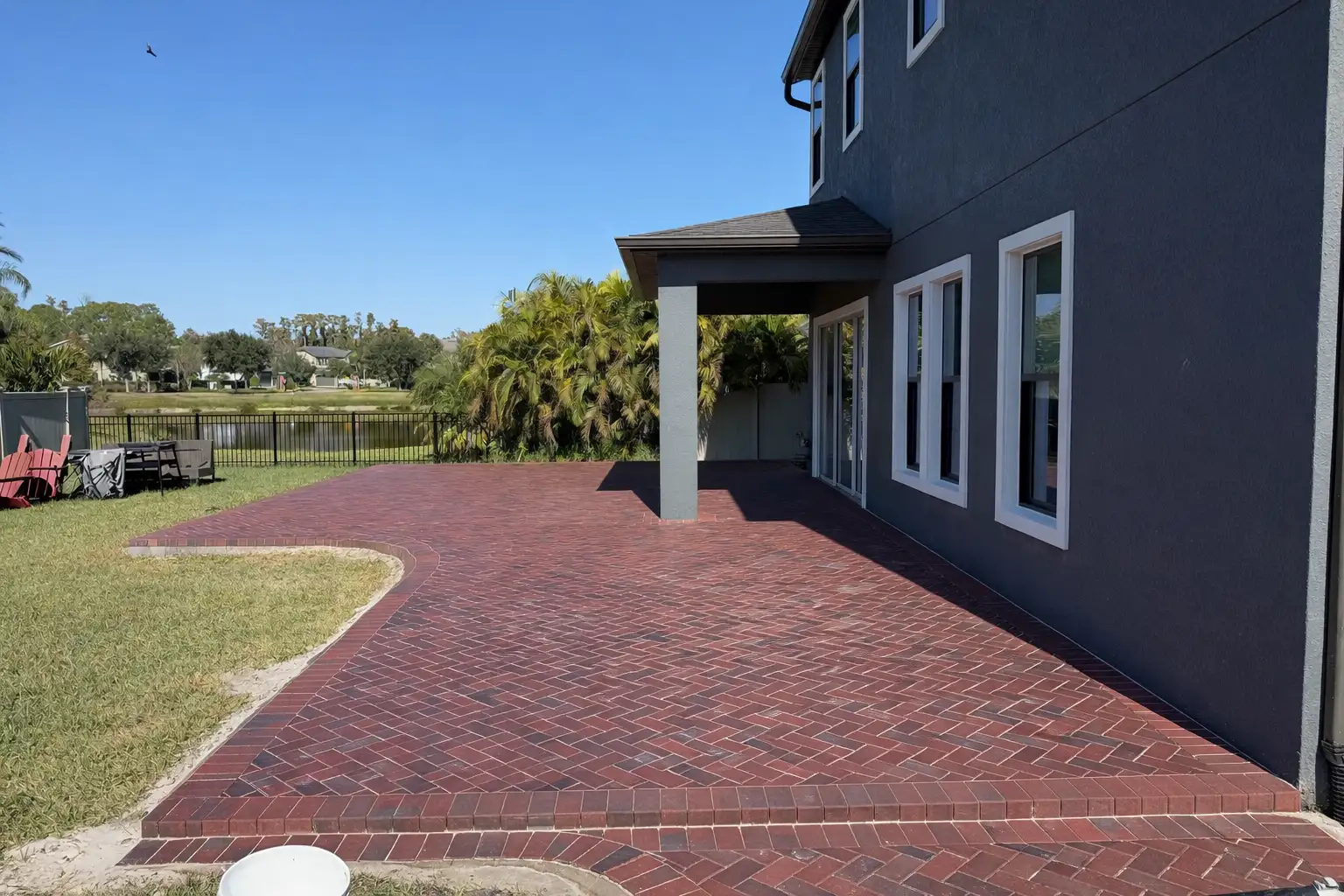Red brick paver patio in herringbone pattern beside grey house with multiple windows and covered porch, under clear blue sky