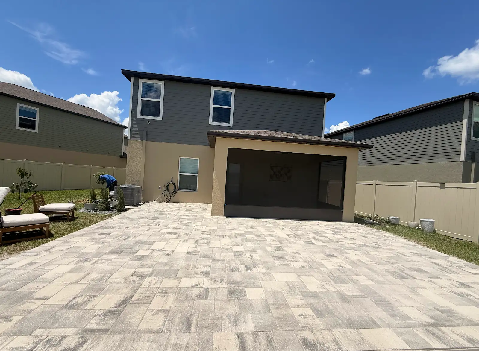 Wide view of a backyard with newly installed light multi-toned paver patio extending from a two-story house with a covered porch and outdoor chairs on grass.