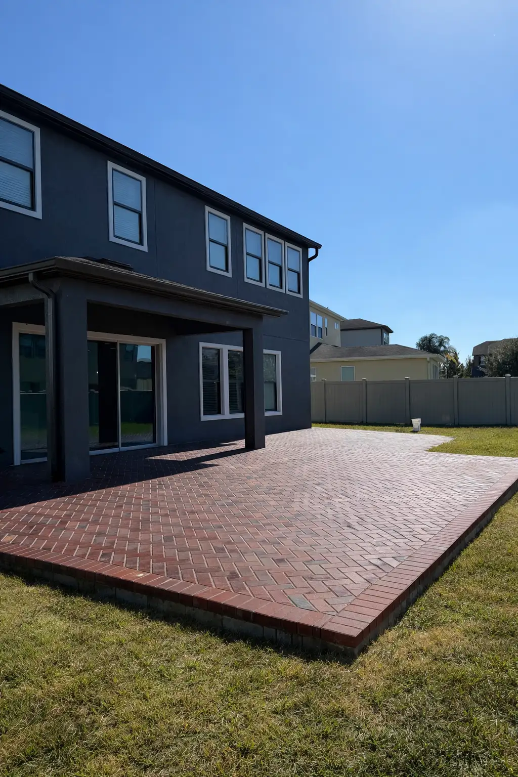 Brick paver patio in herringbone pattern extends from a two-story dark gray house under clear blue sky with green grass bordering the patio.
