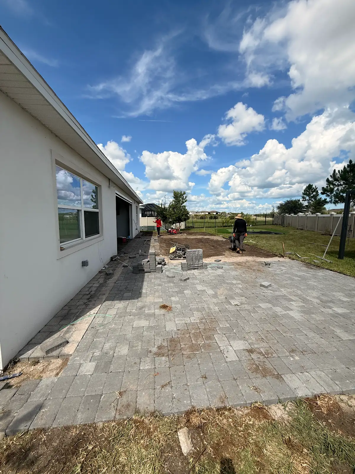 Half-finished grey paver patio extending from a house with workers preparing soil in the backyard under a bright sky with scattered clouds.
