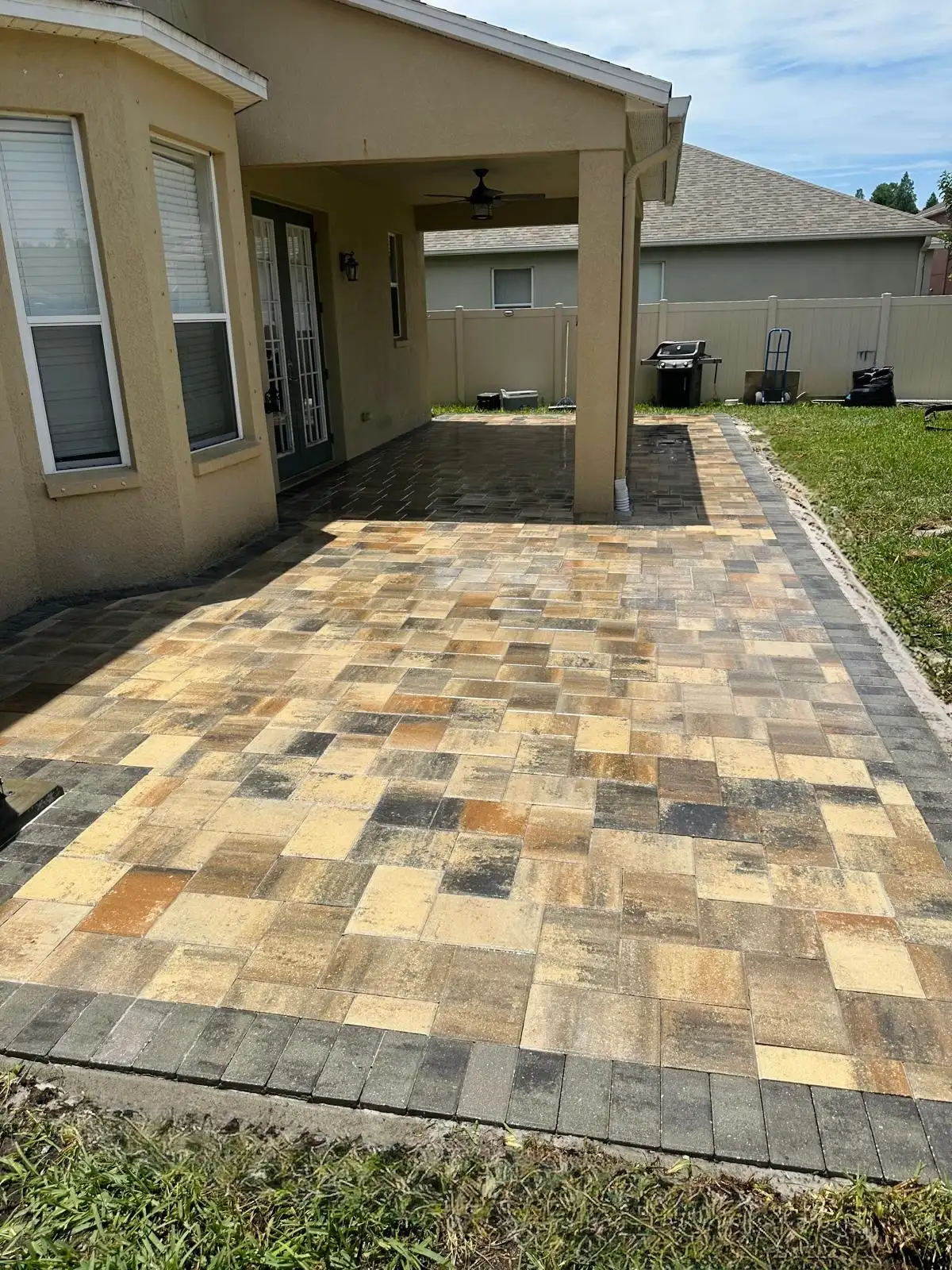 A multi-color paver patio freshly installed beside a covered porch with beige stucco walls and green doors with white blinds in a backyard.