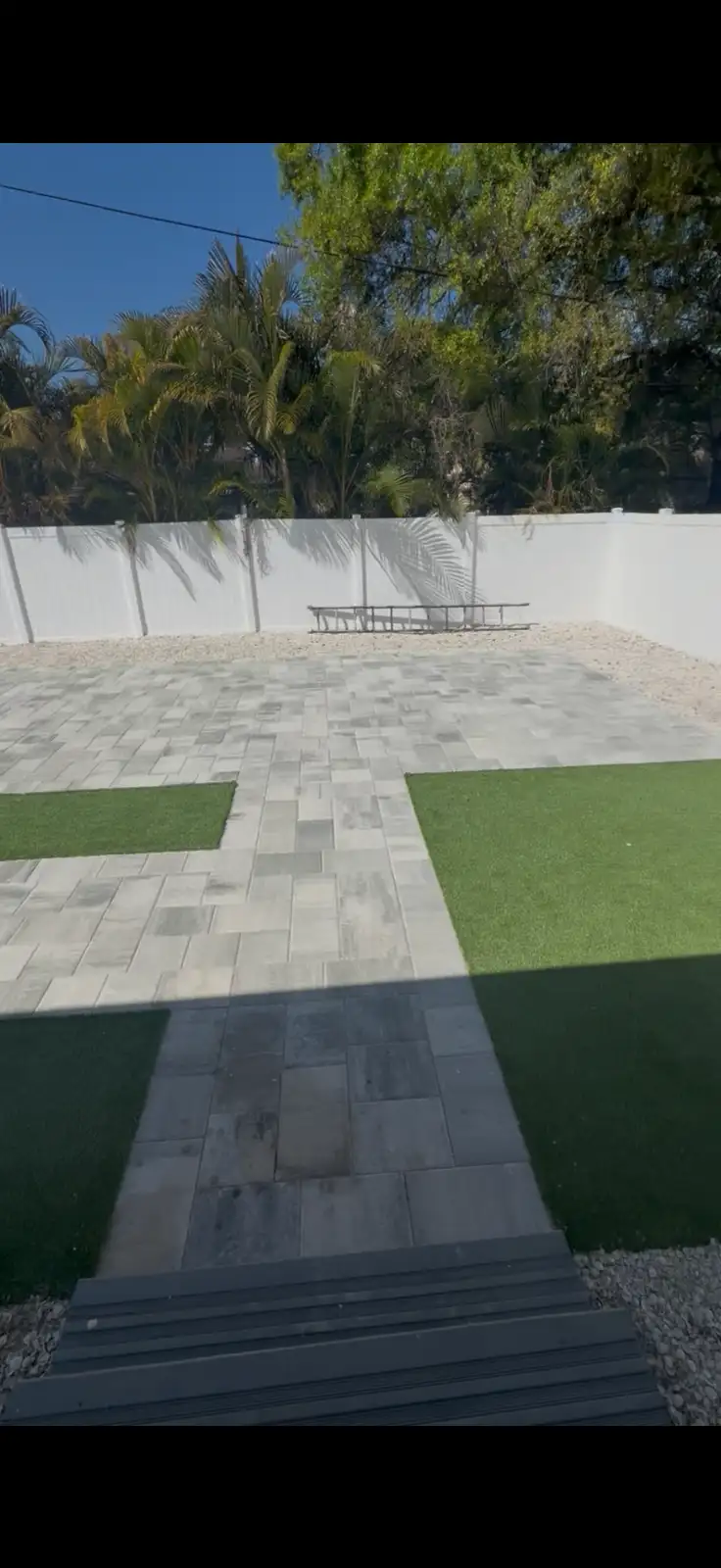 View of a modern paver patio with gray rectangular pavers, artificial turf patches on both sides, white fence backdrop, and tropical trees in the background under clear blue sky.