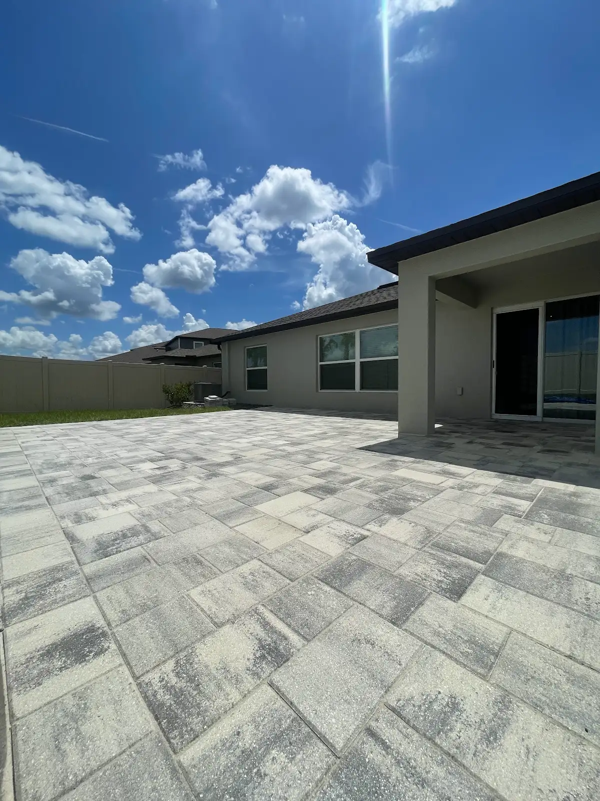 Light gray and beige paver patio outside modern house with large windows and sliding glass door under sunny sky with some clouds