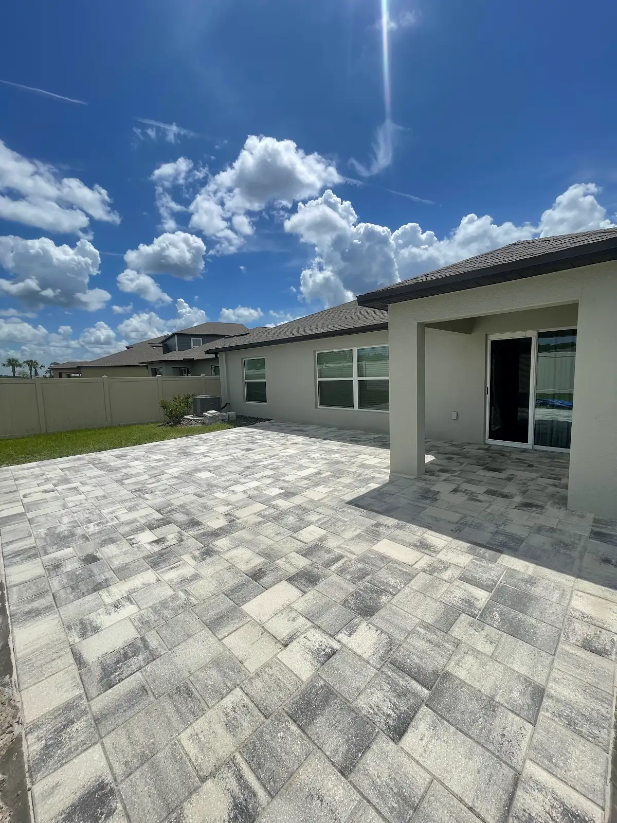 Light gray and white paver patio adjacent to beige house with covered porch under partly cloudy blue sky