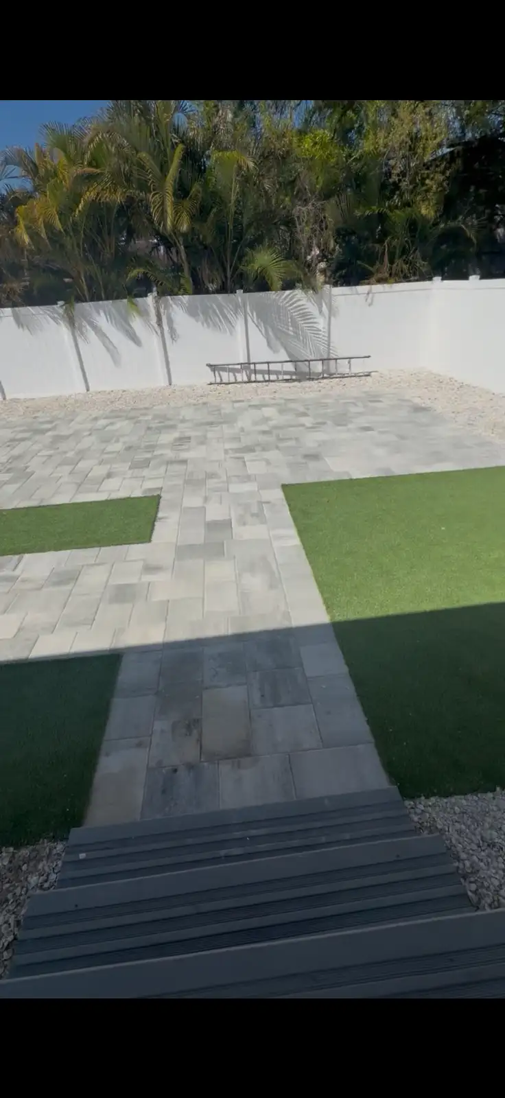 A completed backyard patio with light gray rectangular pavers and green artificial turf patches bordered by a white fence and tropical plants behind it.