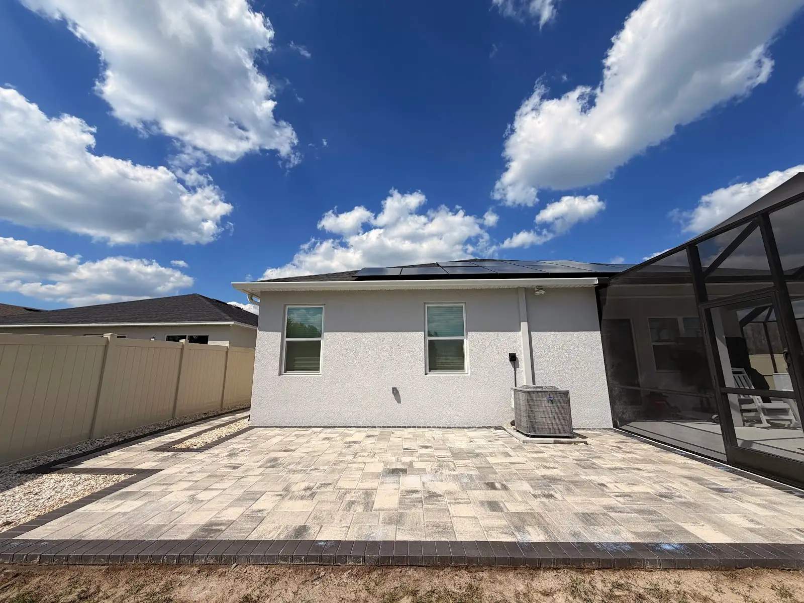 A finished light-colored rectangular paver patio with dark border next to a white house wall with two windows and an air conditioning unit, under a blue sky with clouds.