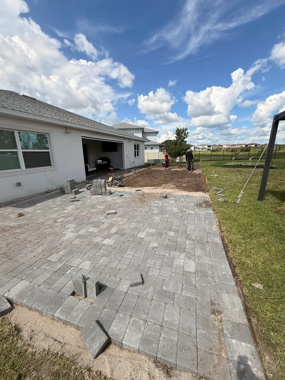 View of a gray paver patio under construction next to a white house, with workers preparing the unfinished section and cloudy sky above