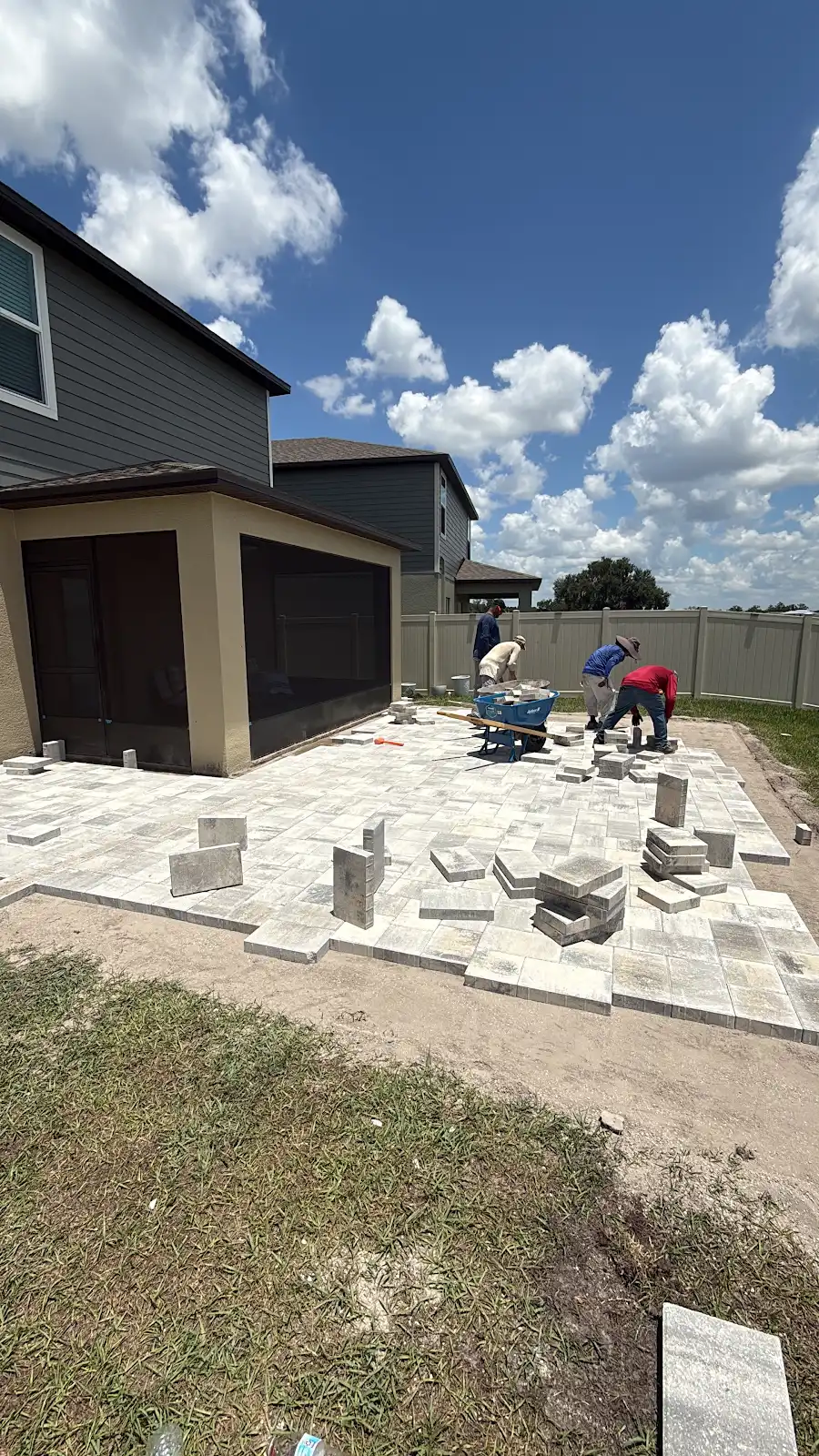 Workers installing light-colored paver patio next to a screened porch attached to a house under blue sky with clouds
