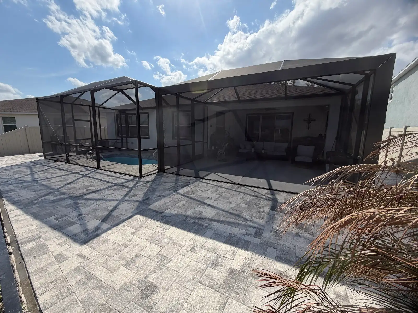 Newly installed light gray rectangular pattern paver pool deck around a screened pool enclosure attached to a residential house, under partly cloudy sky.