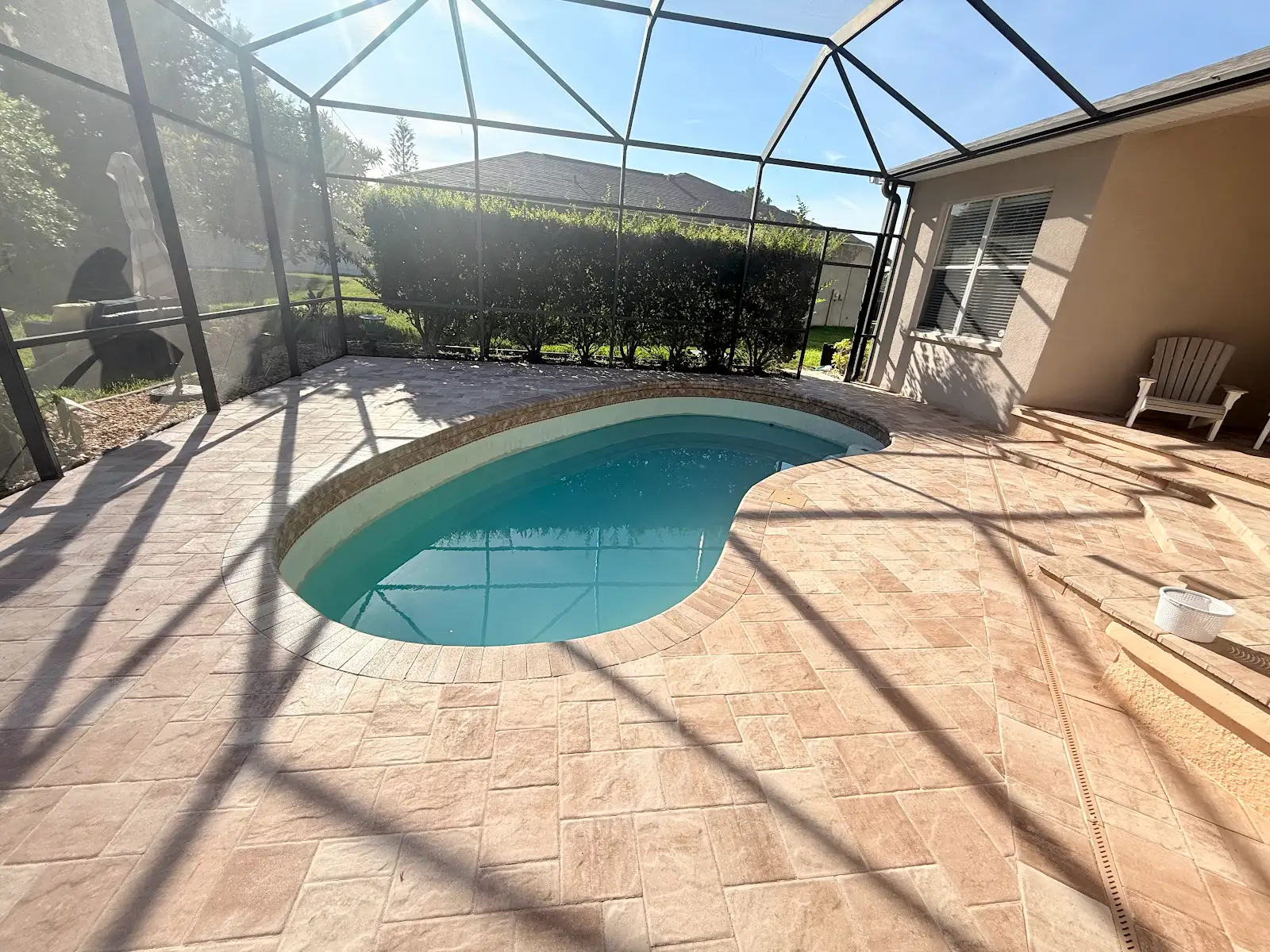 Kidney-shaped pool with light-colored paver pool deck under a screened enclosure next to house with steps and chair.