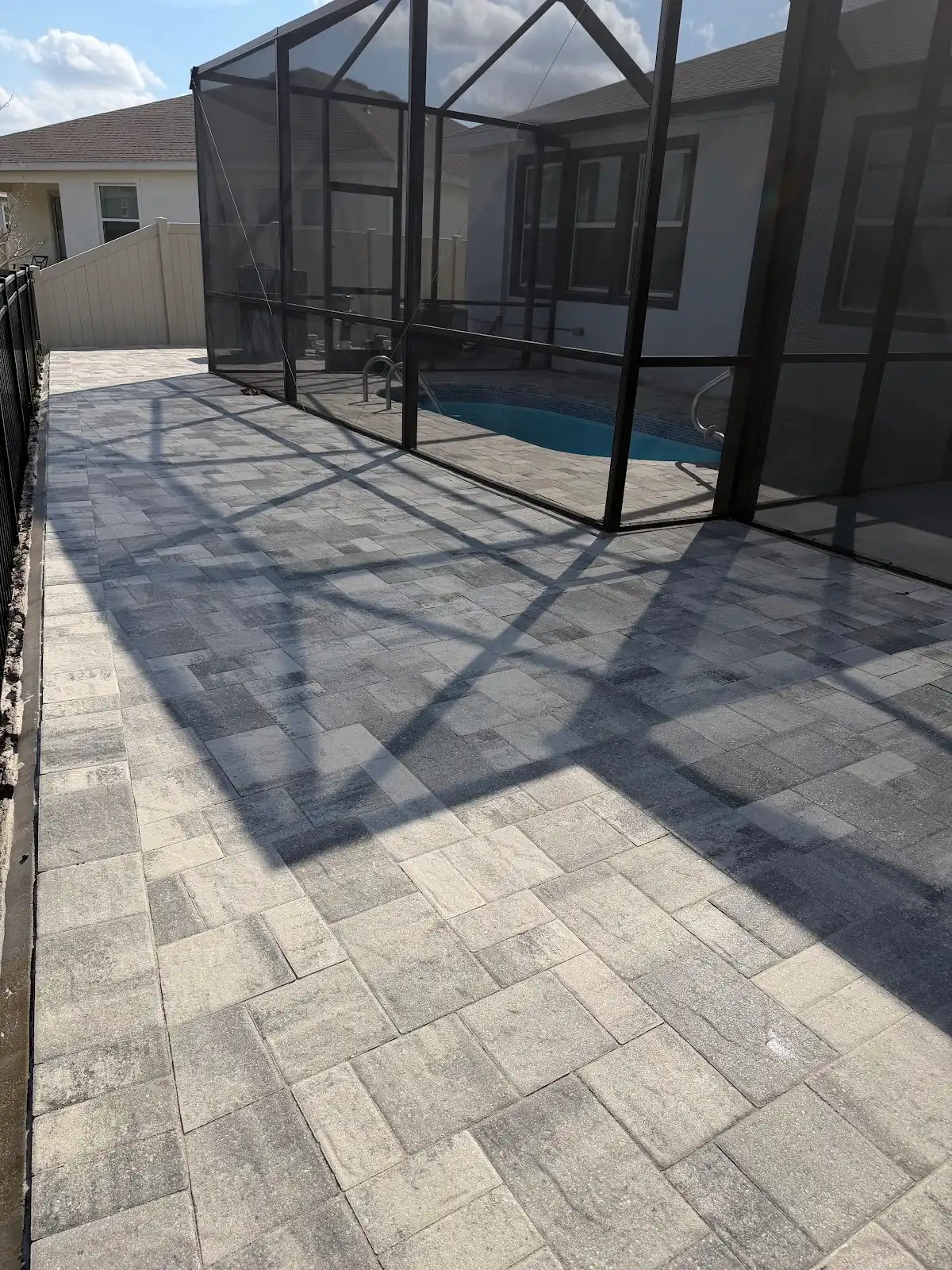 Grey and beige patterned paver pool deck around a screened pool enclosure adjacent to a house under blue sky
